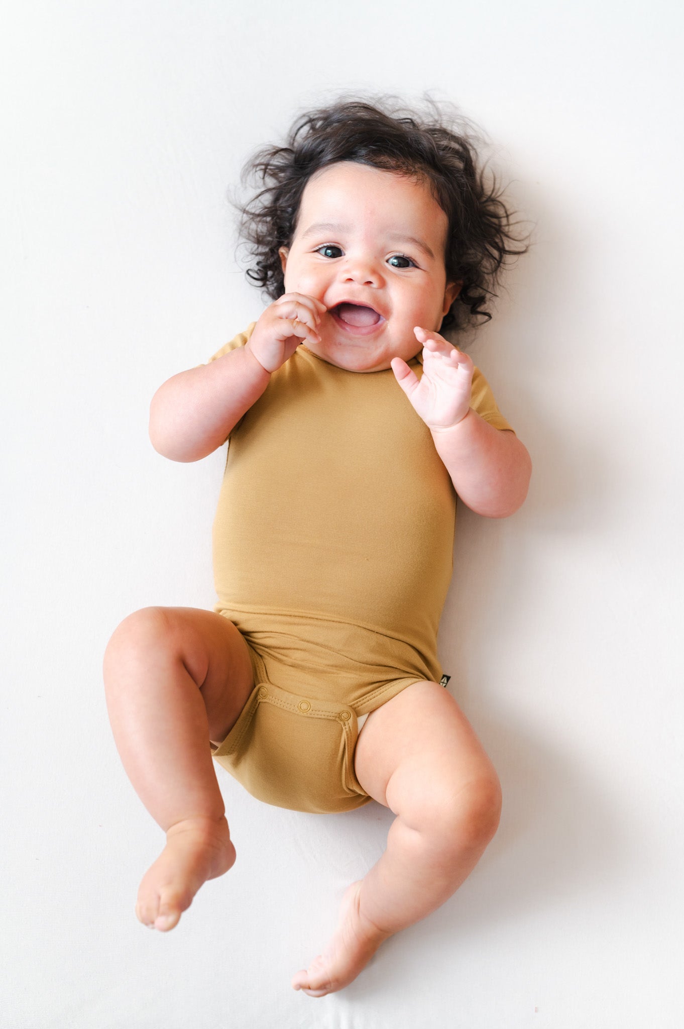 A smiling baby lying on their back wearing a mustard yellow short-sleeve bodysuit, photographed against a clean white background.