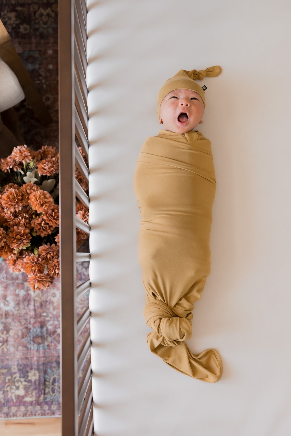 A newborn baby swaddled in a mustard yellow fabric wrap and matching hat, lying on a white baby crib mattress next to a wooden crib frame and dried flowers.