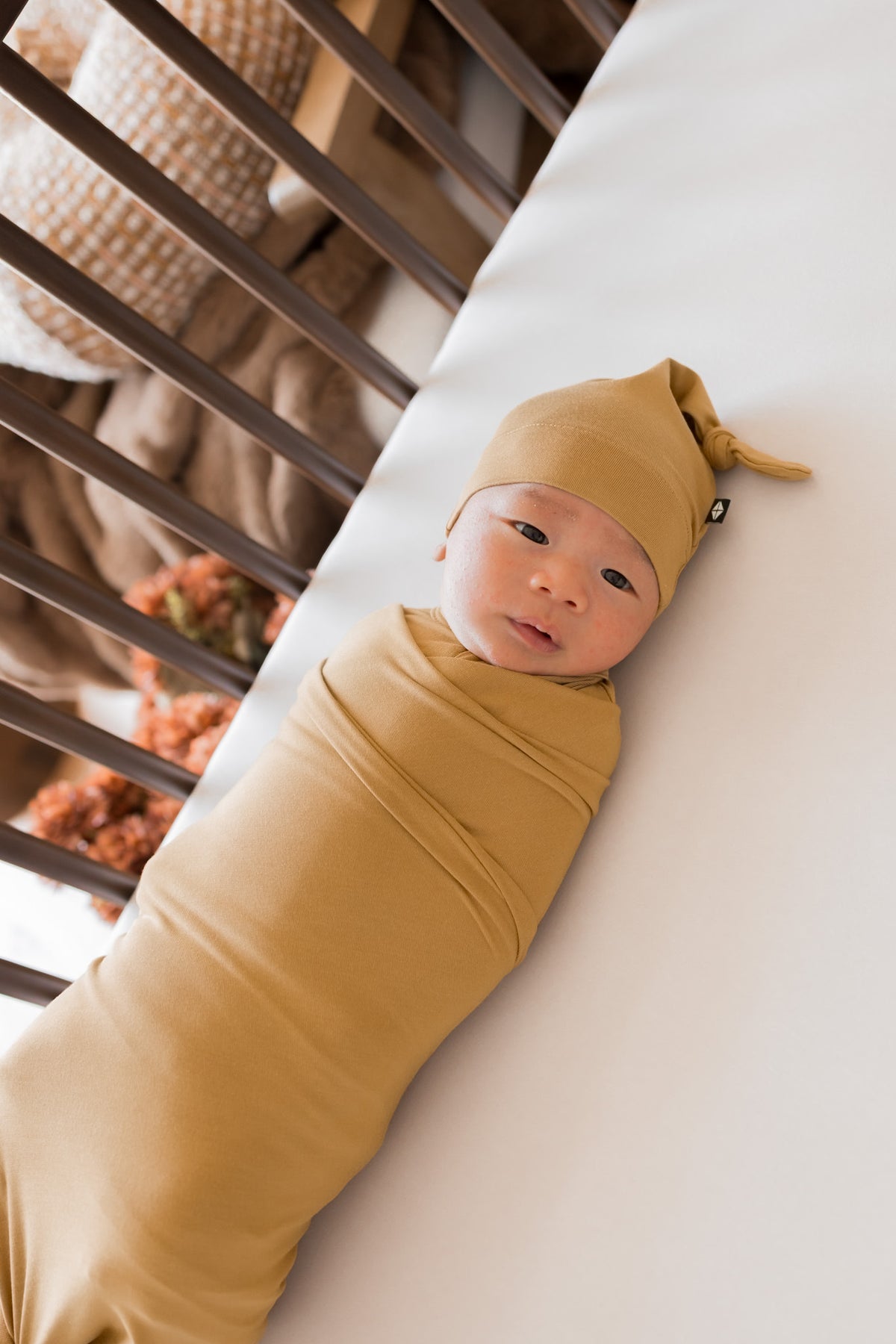 A baby swaddled in mustard yellow fabric and wearing a matching knotted beanie hat, lying on a white mattress next to a dark wooden crib.