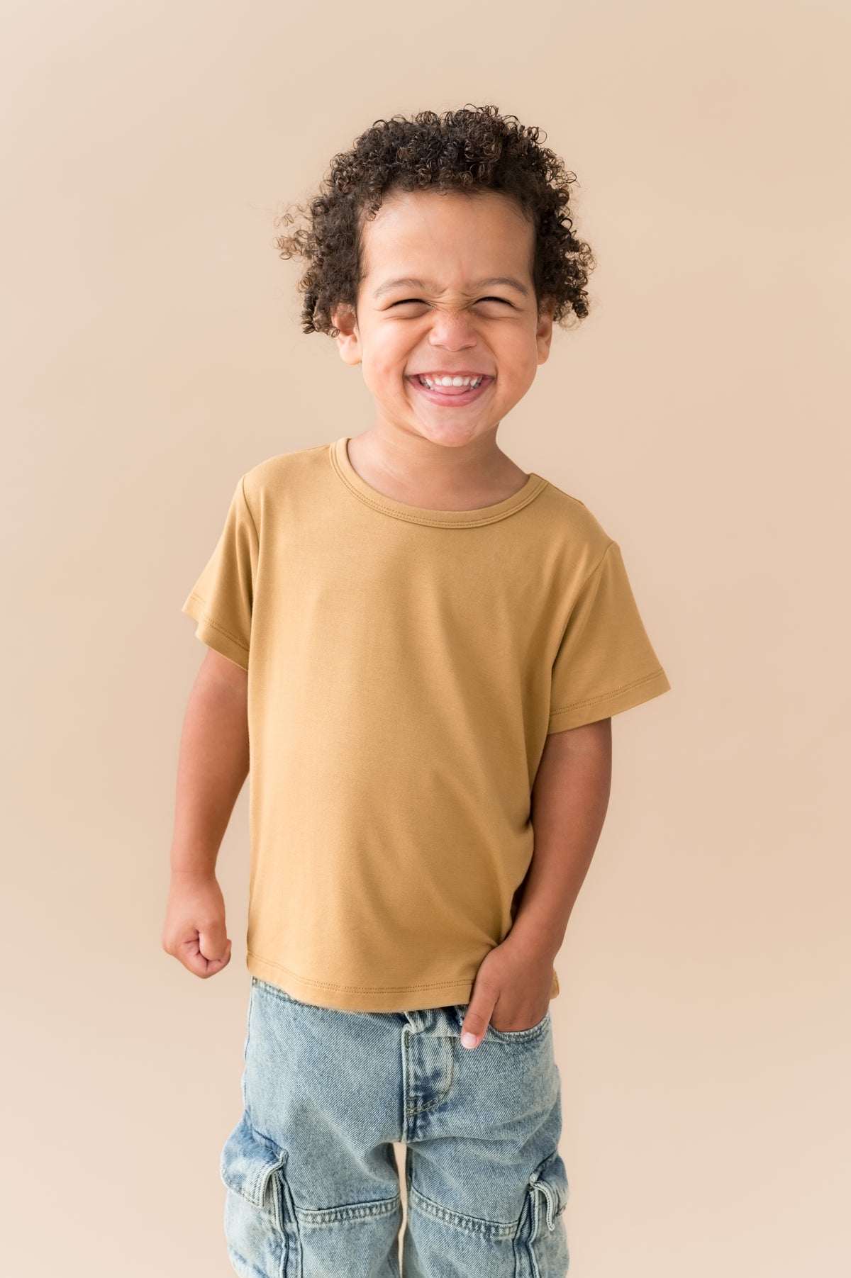 Close up of Boy wearing Toddler Basic Tee in Ochre over blue jeans on a beige background