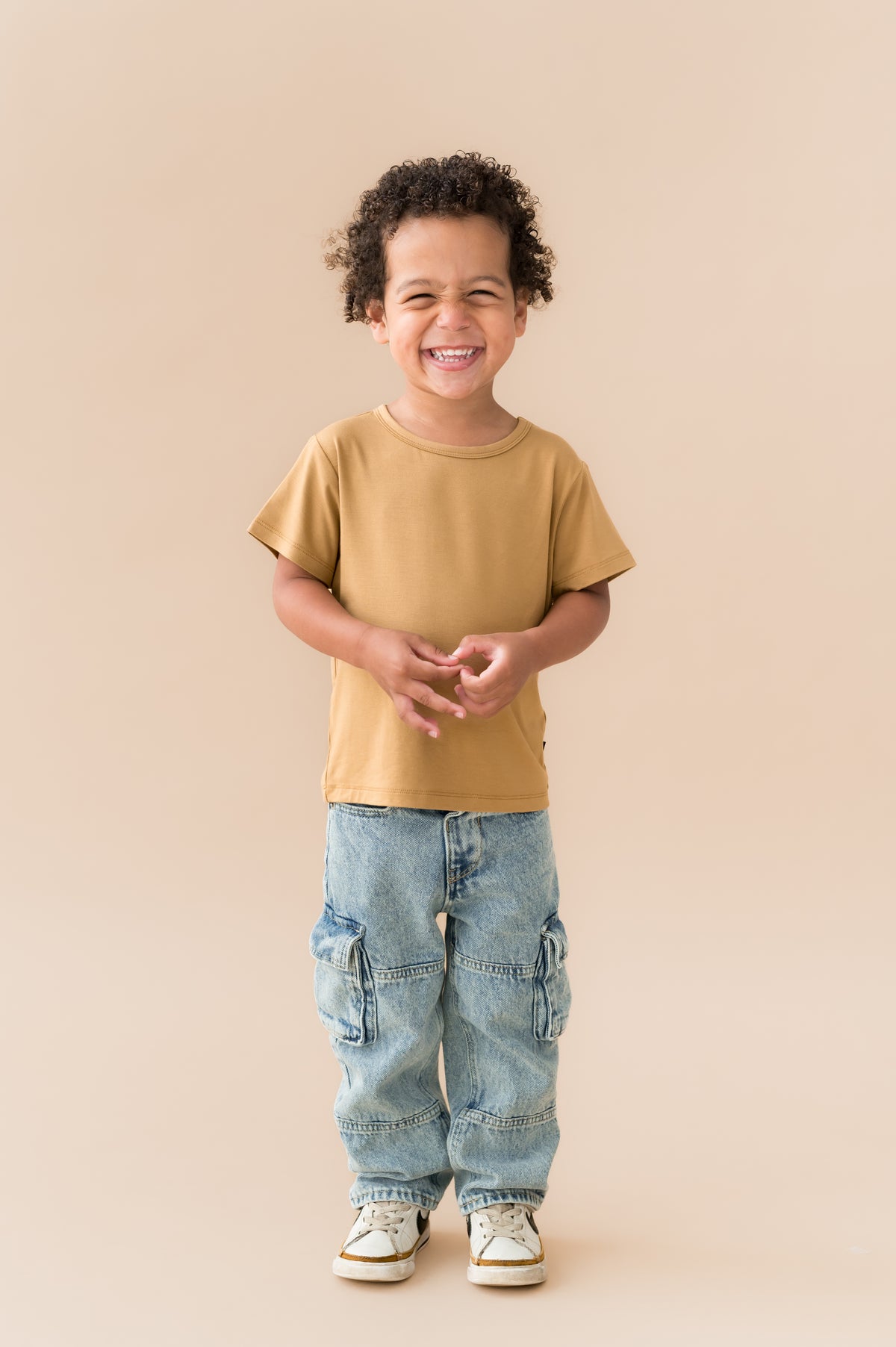 Boy smiling in Toddler Basic Tee in Ochre over blue jeans on a beige background