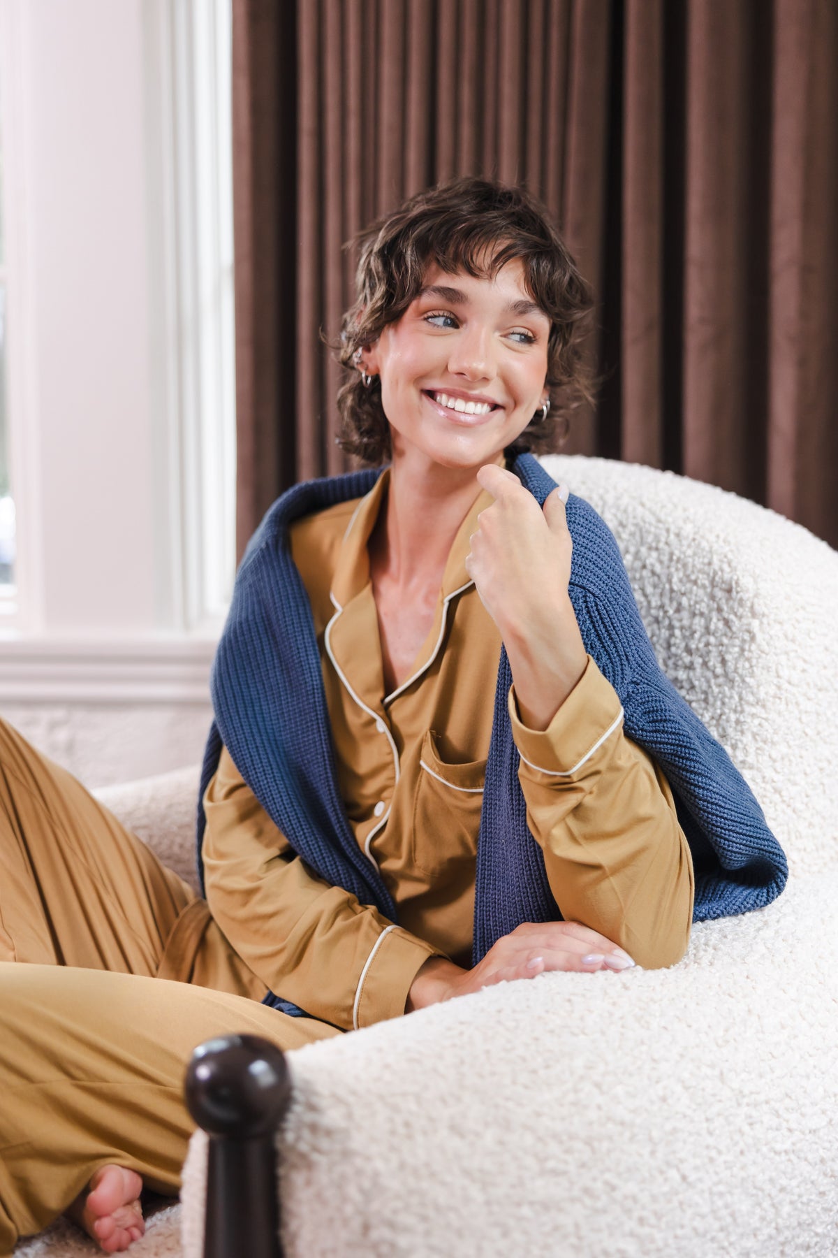 Modal smiling in Long-Sleeved Women's Pajama Set in Ochre with blue sweater over her shoulders sitting on a sofa
