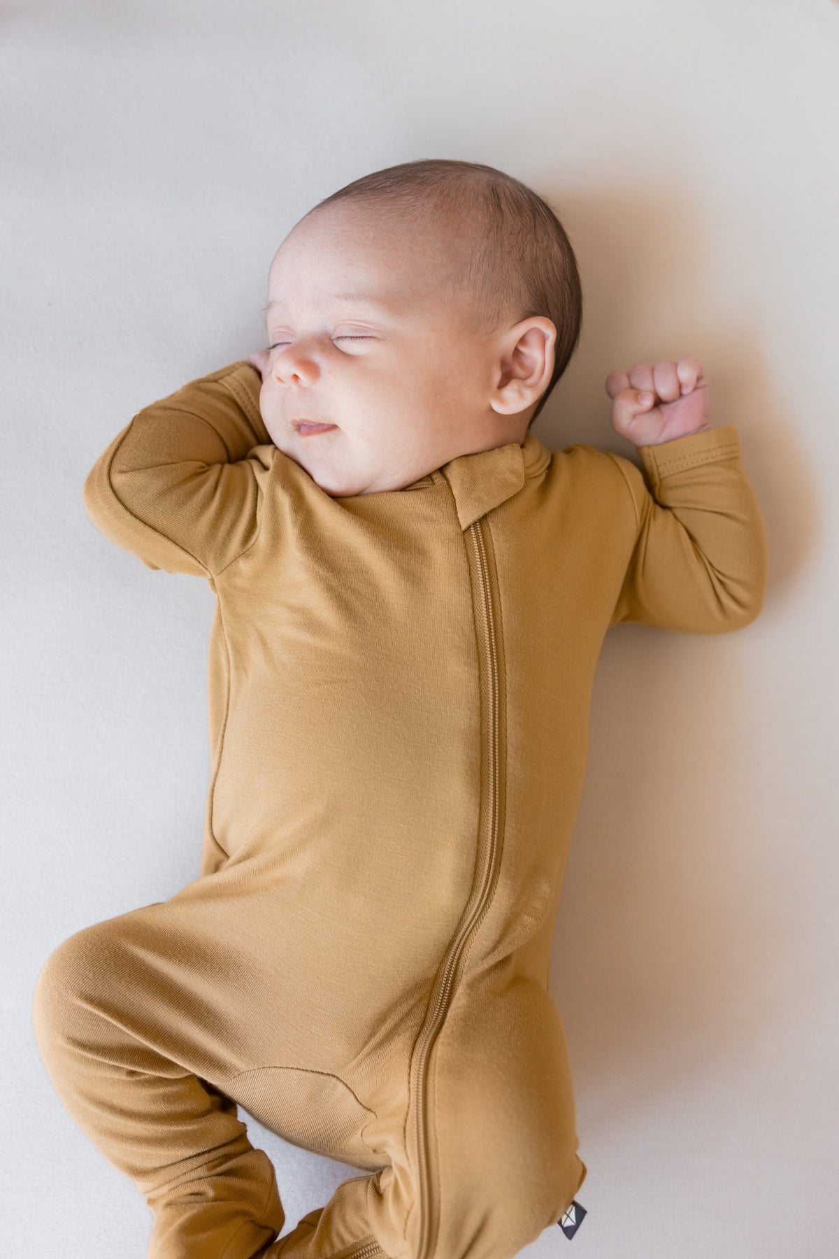 A sleeping newborn baby wearing a mustard yellow full-body zippered onesie, lying on a light neutral background.