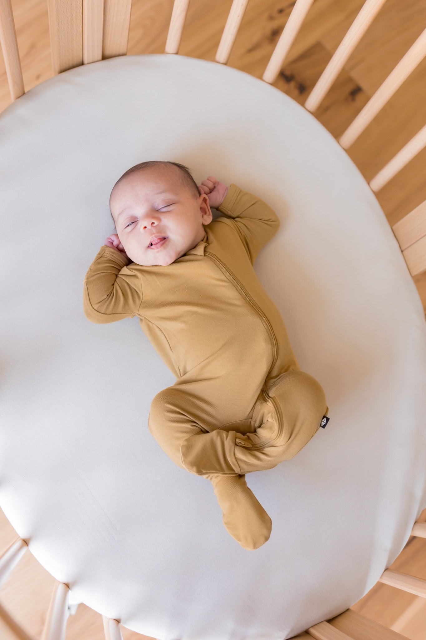 A sleeping newborn baby wearing a mustard yellow long-sleeve footed sleeper, lying on a white mattress inside a wooden crib.