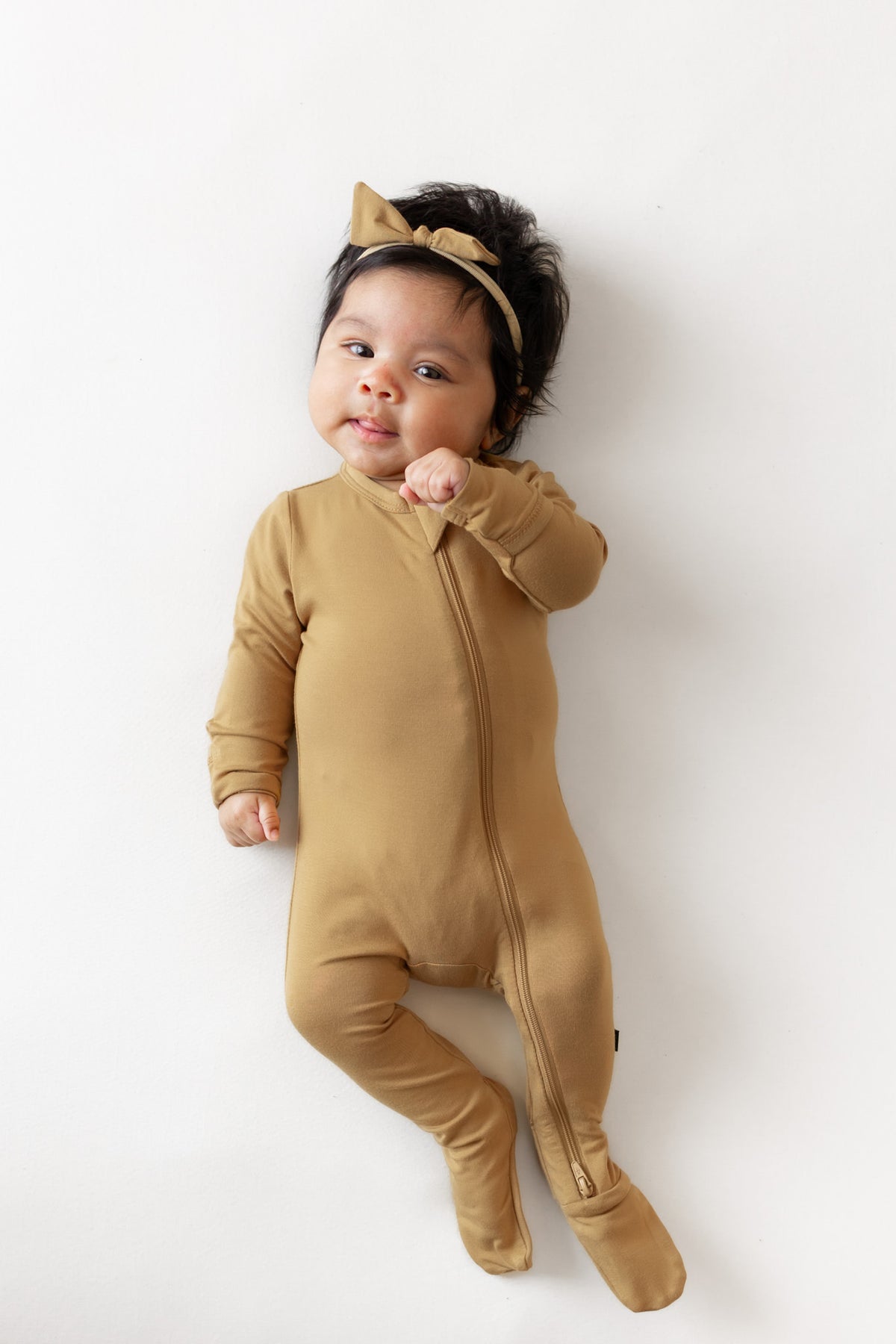 A baby lying on a white background wearing a mustard footed zip sleeper and a matching mustard headband with a bow.