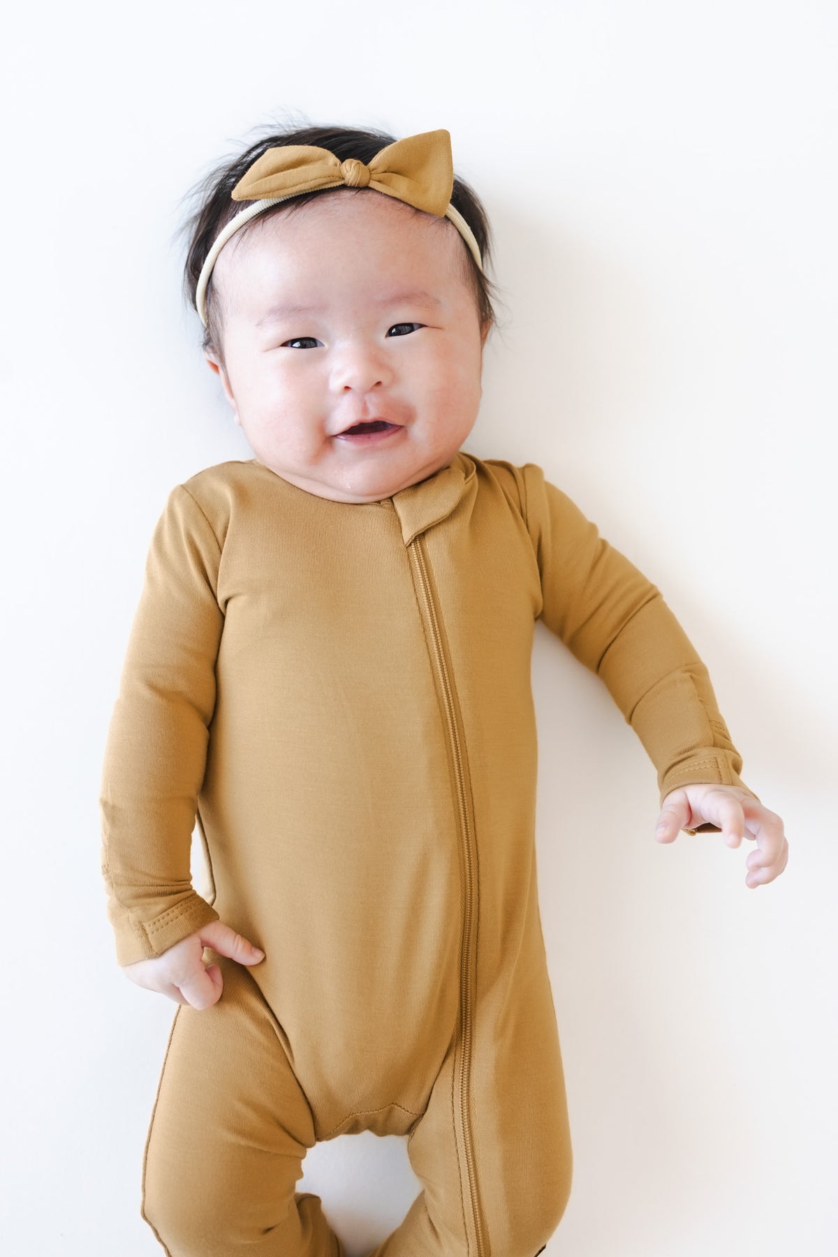 A baby wearing a mustard yellow footless one-piece romper with Tiny Nylon Bow in Ochre, lying on a plain white background.