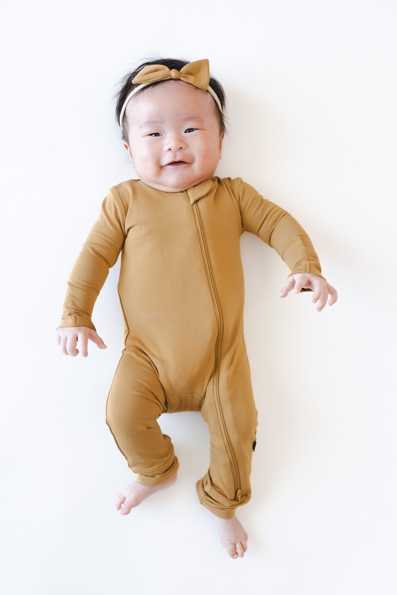 A baby lying on their back wearing a mustard yellow long-sleeve footless romper with a matching bow headband, photographed against a clean white background.