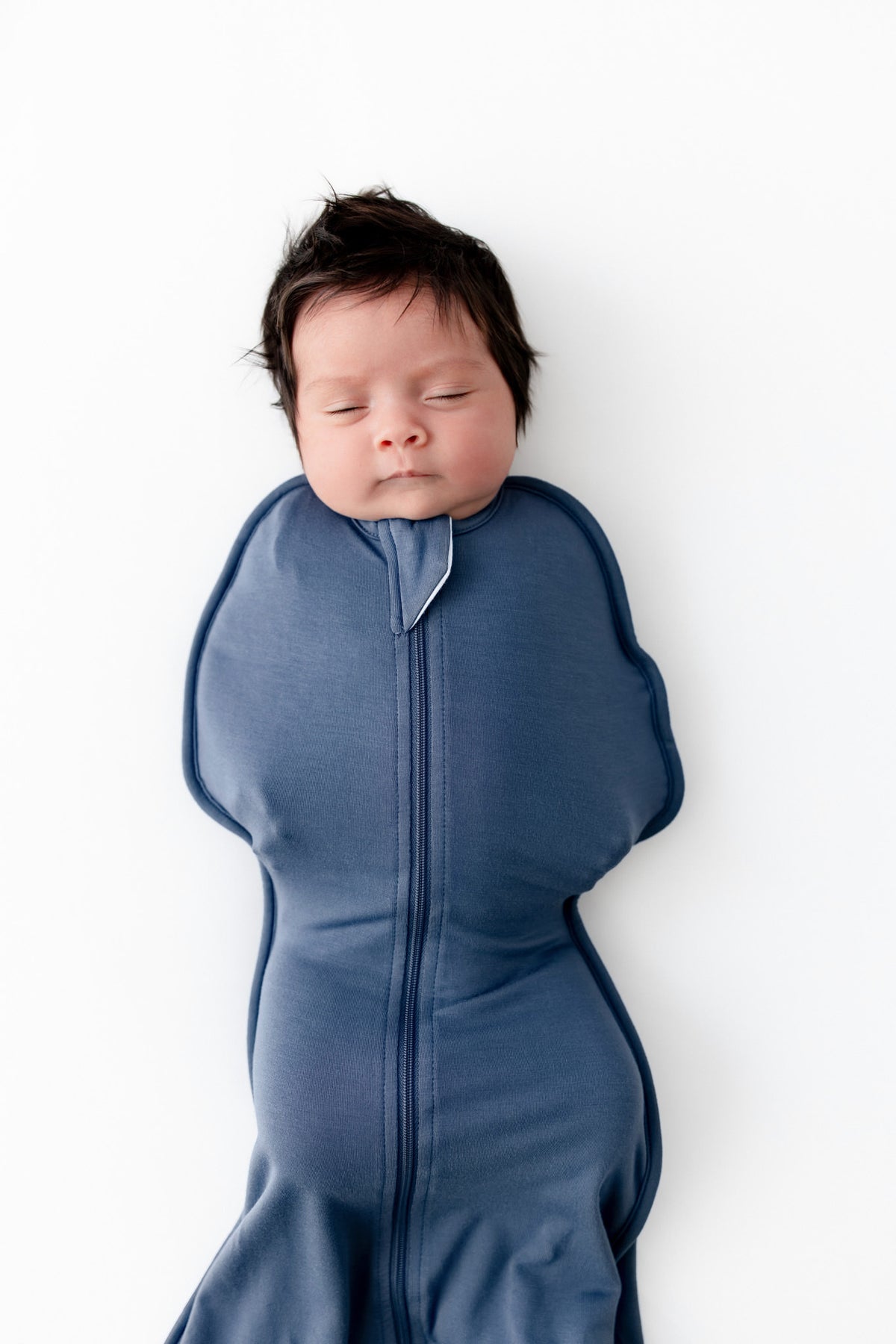 A sleeping newborn baby swaddled in a slate blue swaddle bag, lying on a clean white background.