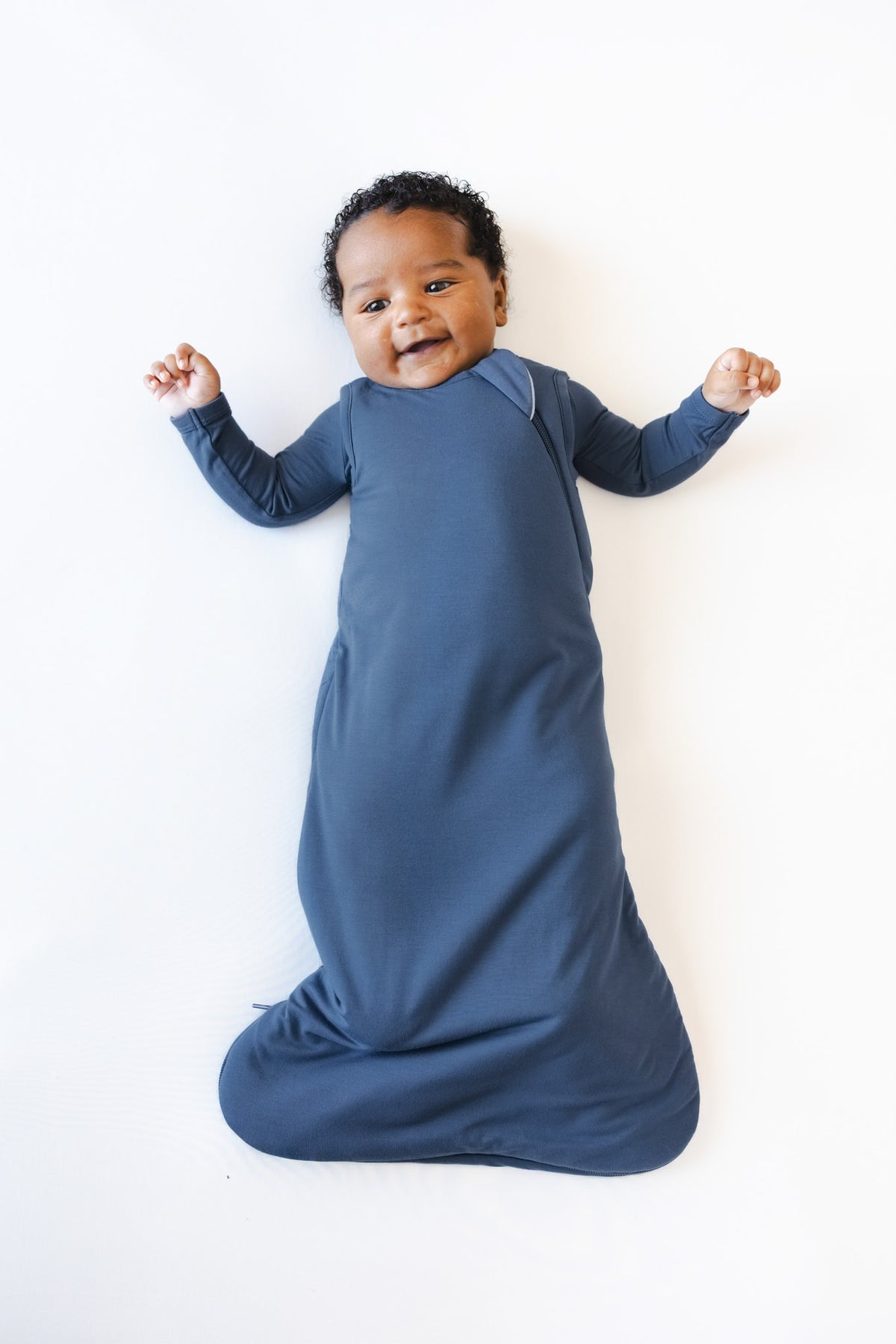 A baby lying on their back wearing a slate 1.0 TOG sleep sack against a clean white background.