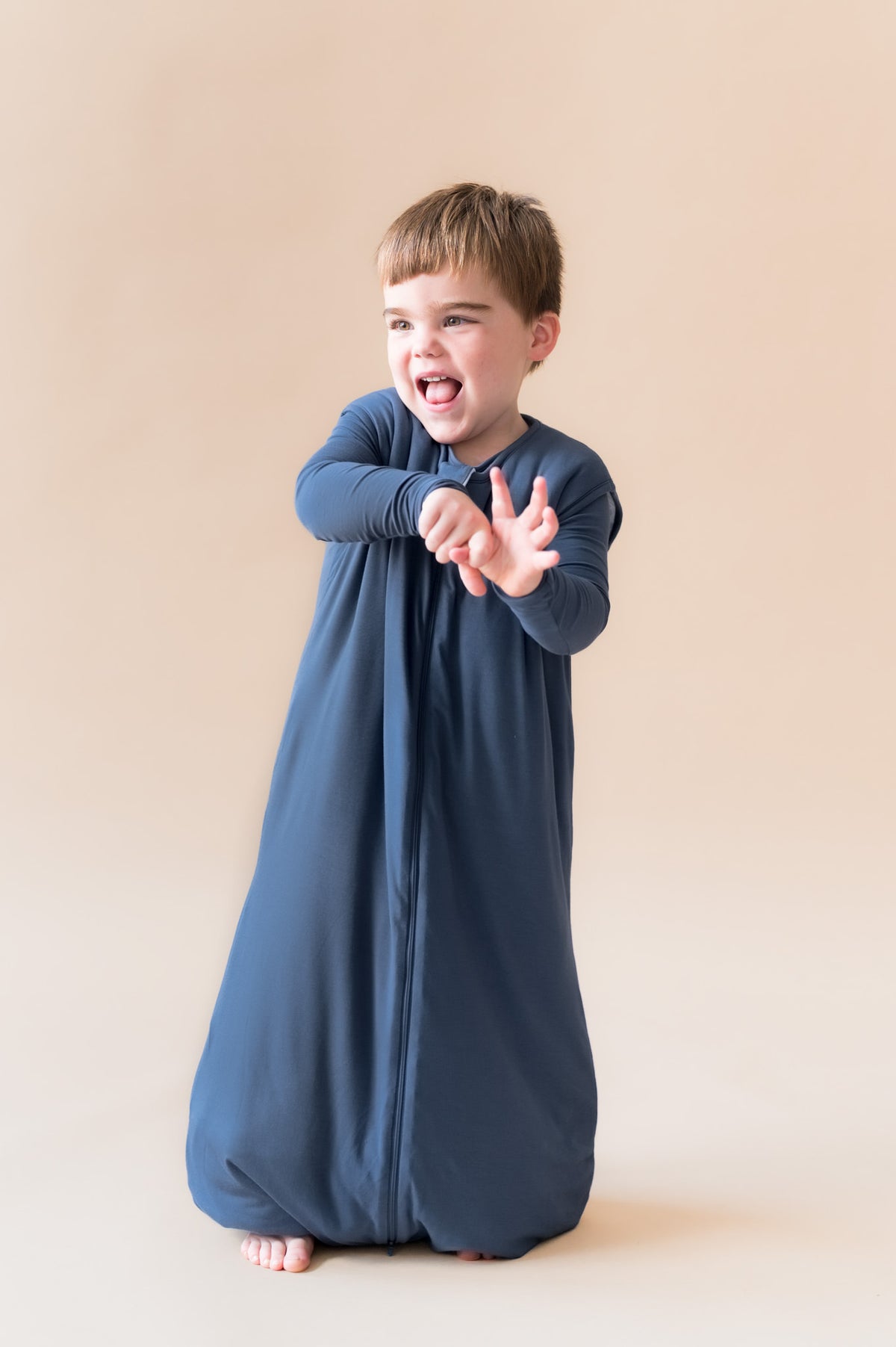 A toddler wearing a navy blue long-sleeve sleep sack with a front zipper, standing against a solid beige studio background.