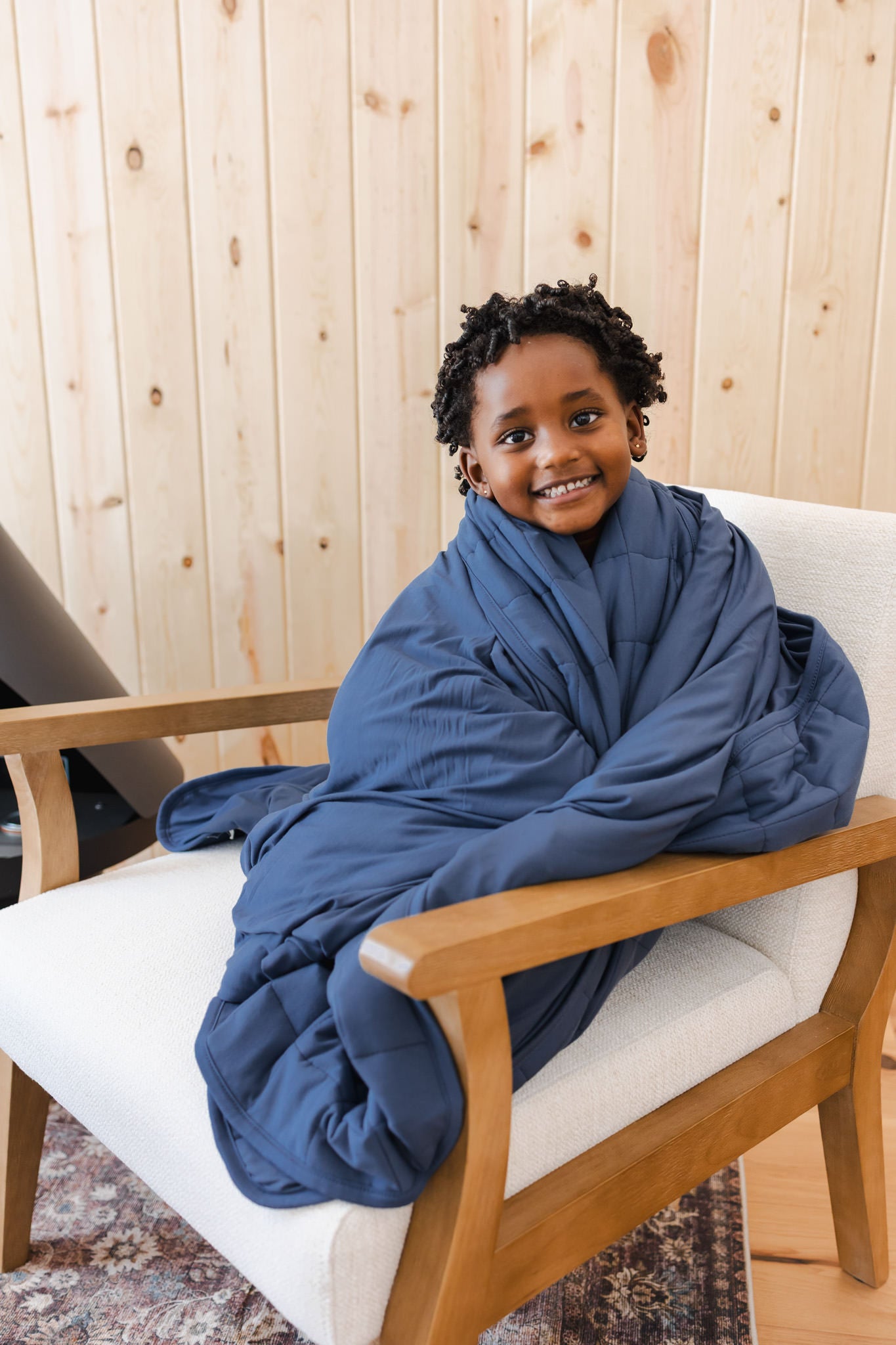 Boy smiling with Youth Blanket in Oxford 2.5 wrapped around him sitting on a white sofa in a room