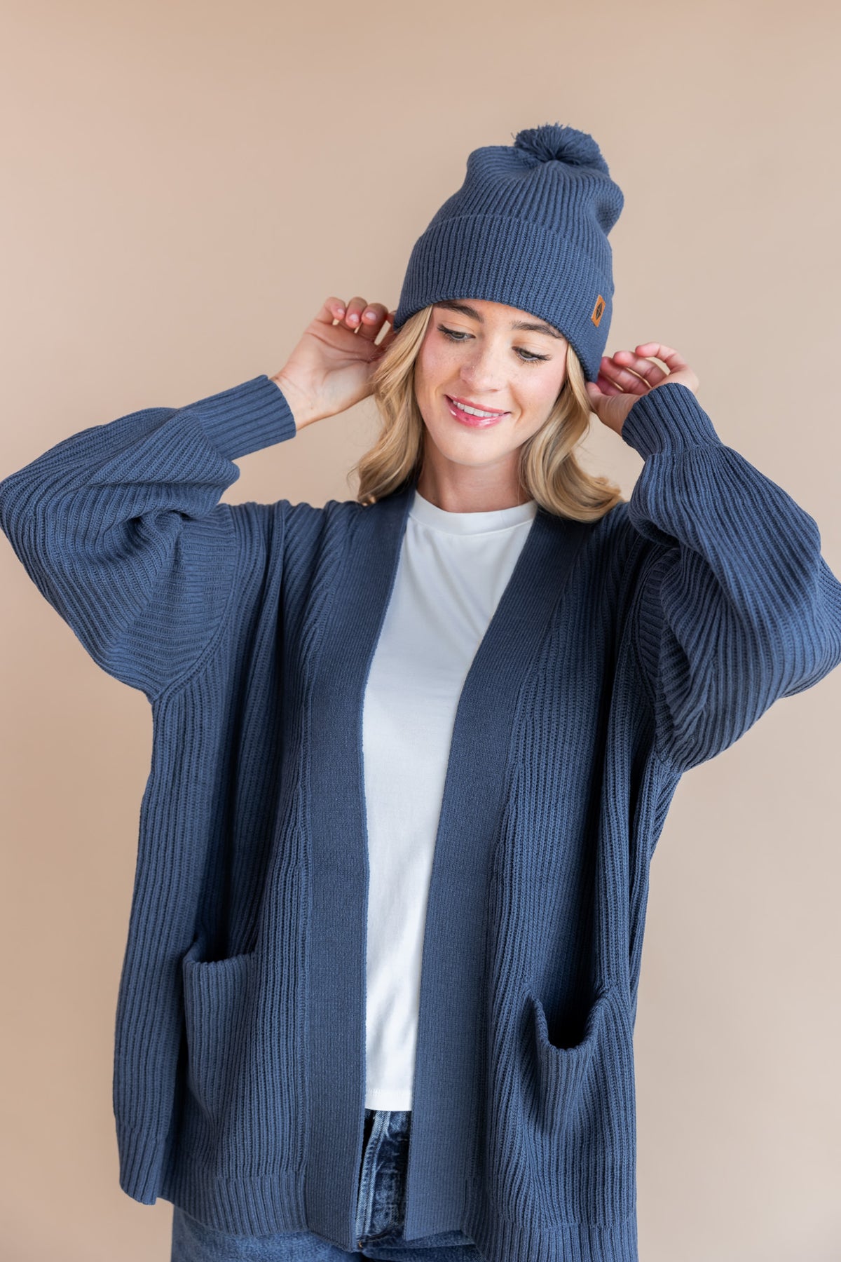 Modal posing in Chunky Knit Adult Pom in Oxford over Chunky knit cardigan in oxford and white T-shirt on a beige background