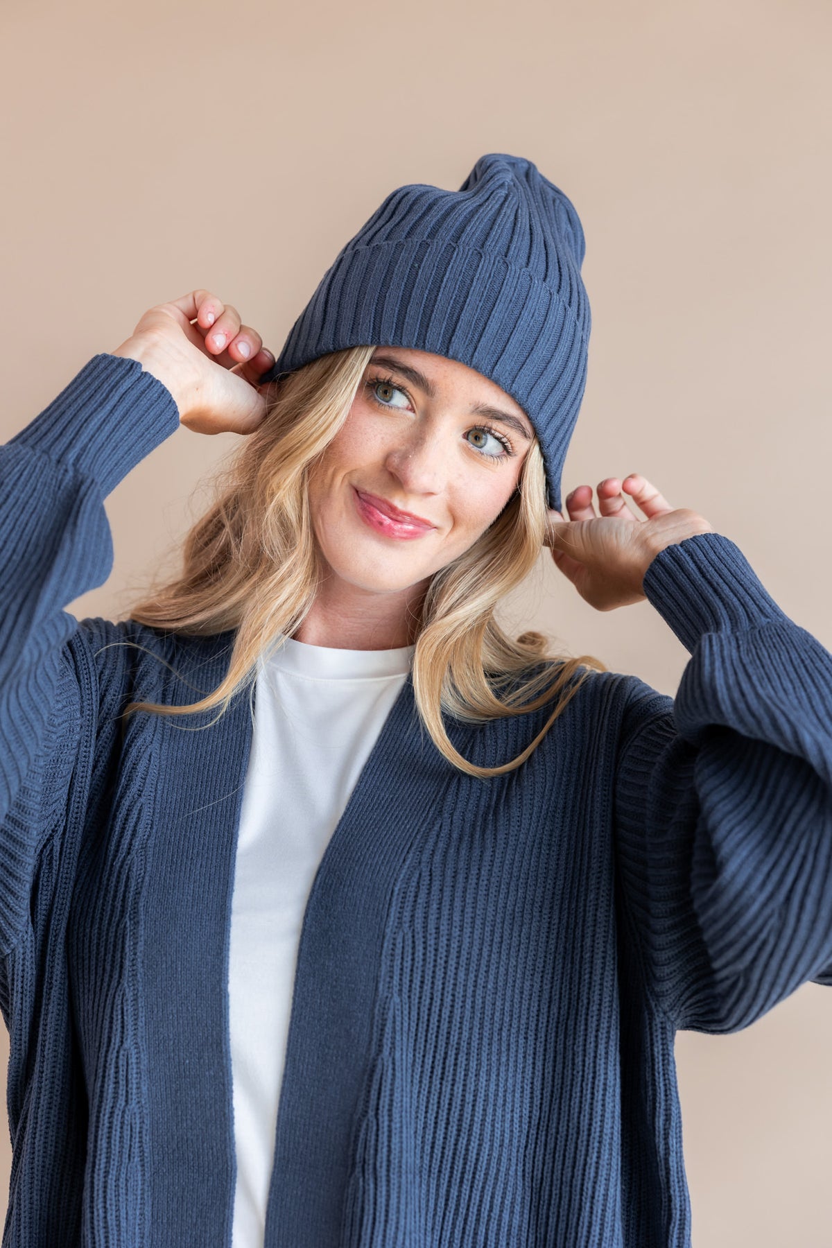 Modal holding Chunky Knit Adult Ribbed Beanie in Oxford over white T-shirt and Chunky knit cardigan in oxford on a beige background