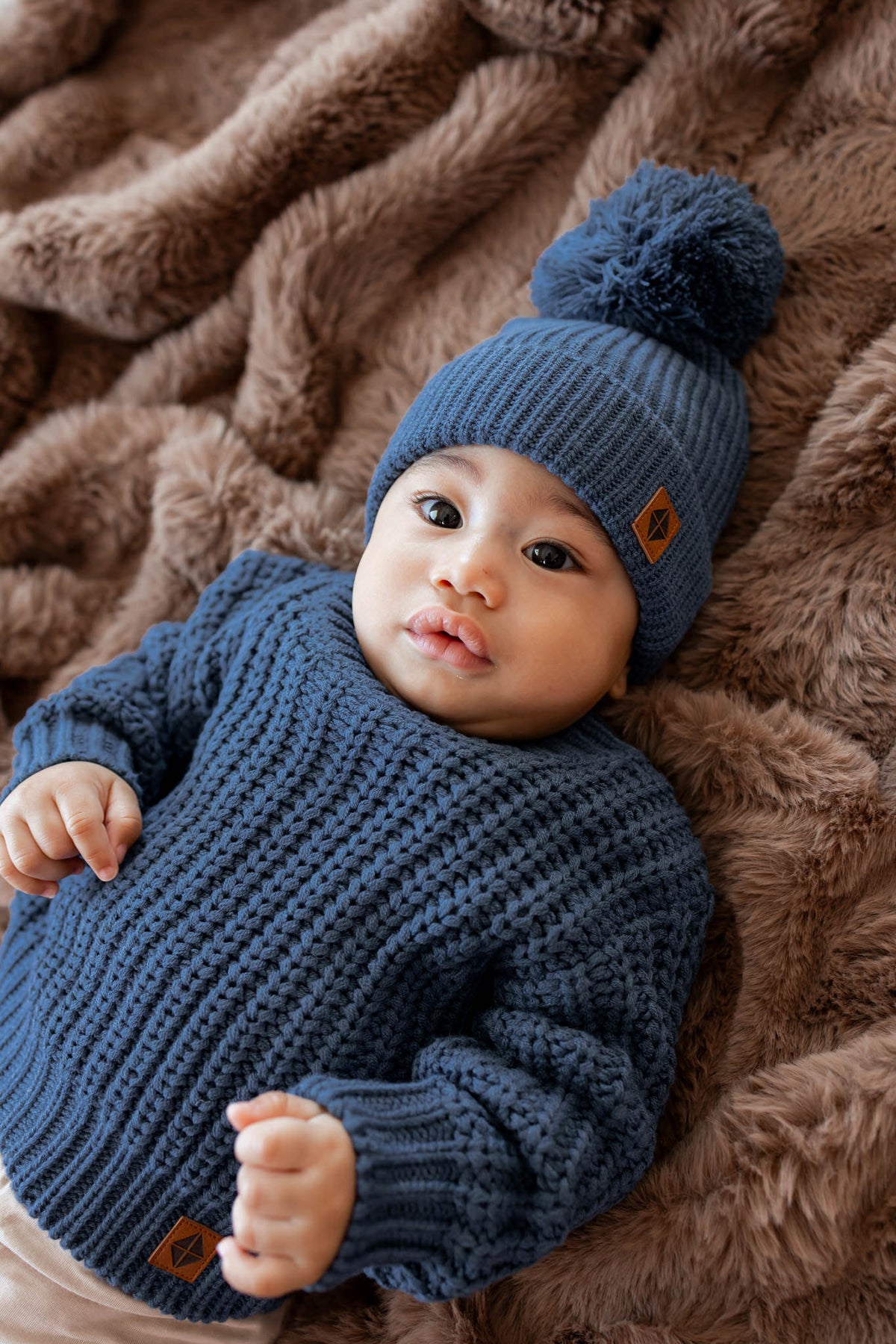 A baby wearing a blue knit sweater and matching blue pom-pom beanie lies on a brown faux fur blanket. Small brown brand tabs are visible on both garments.