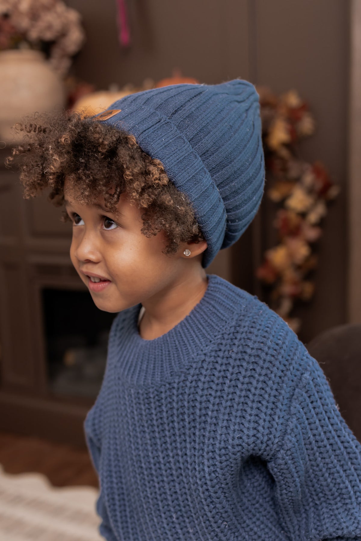A young child wearing a blue ribbed knit beanie and a matching blue textured knit sweater, shown in a warm indoor setting.