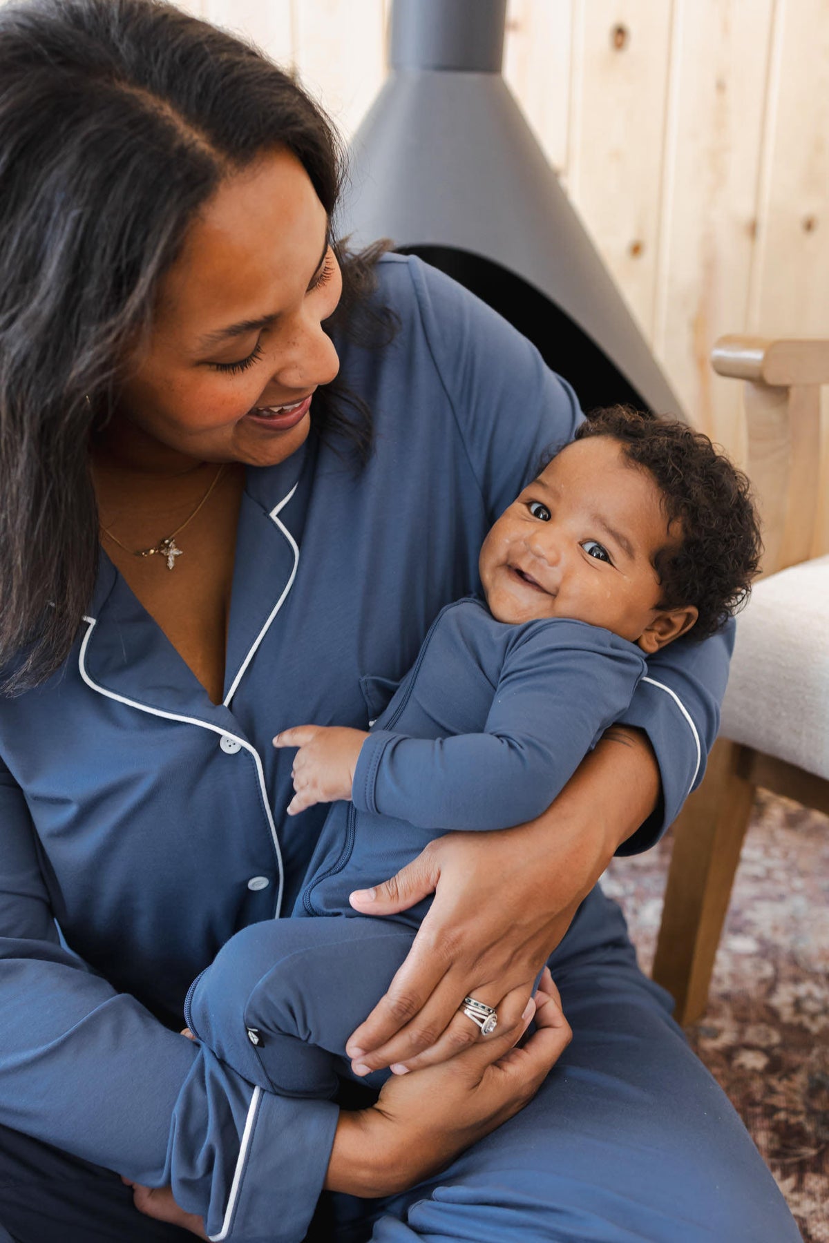 A woman and a baby wearing matching slate blue pajamas with white piping, sitting together in a cozy indoor setting.