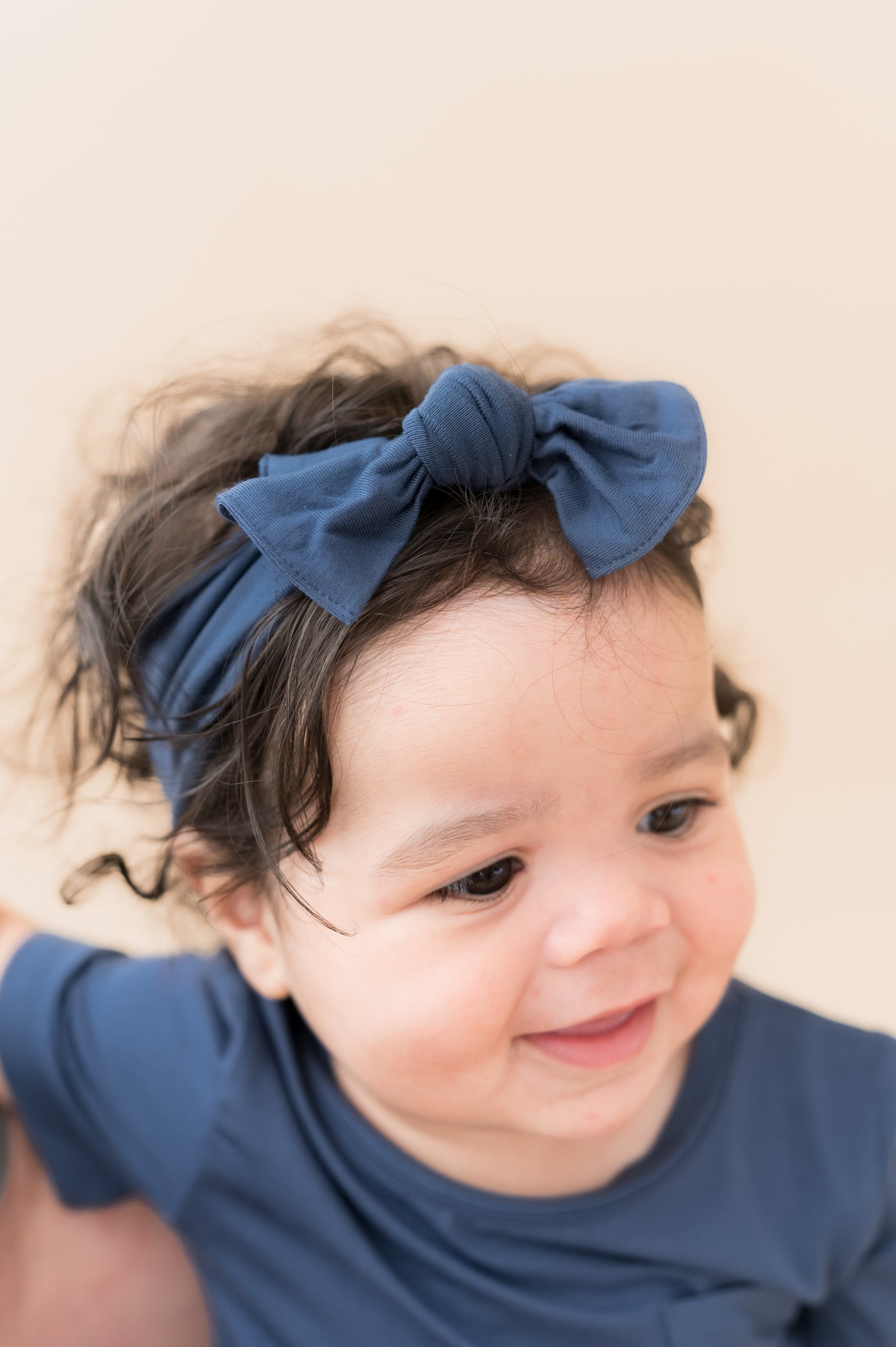 Baby wearing Knotted Bow Headband in Oxford on head over a beige background