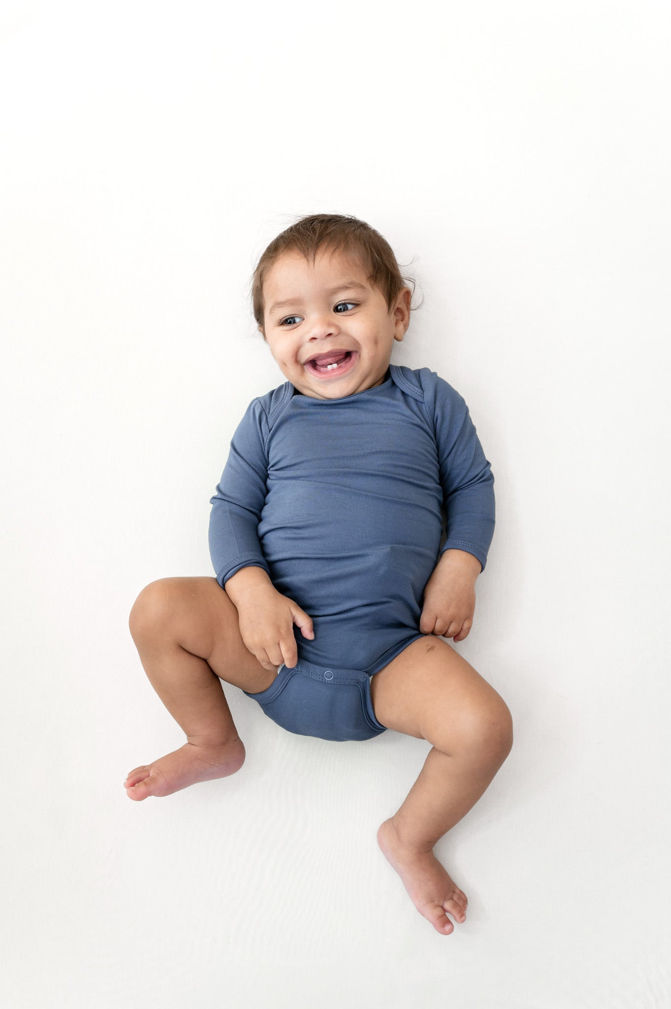 A smiling baby sitting on a white background, wearing a dusty slate blue long-sleeve bodysuit.