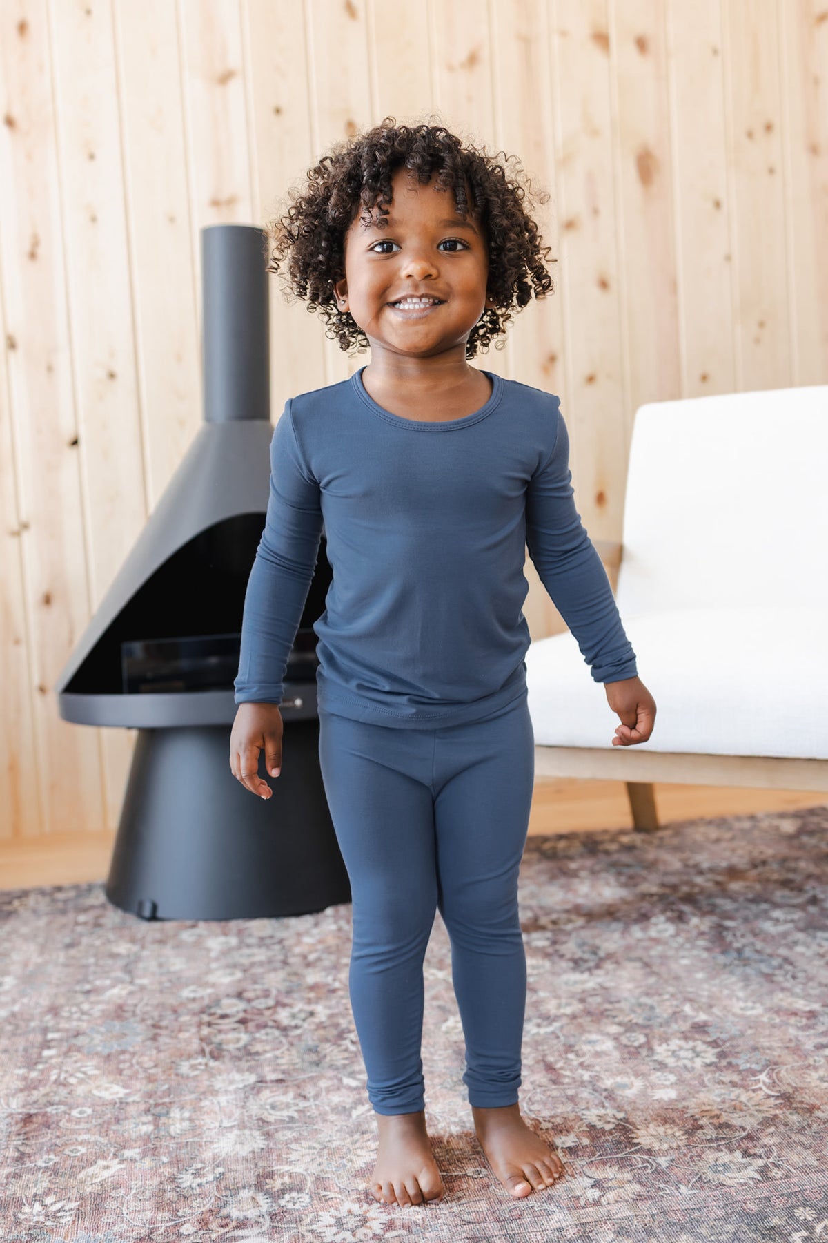 A smiling young child wearing a matching long-sleeve pajama set in a muted slate blue-gray color. The background features a light wood-paneled wall, a modern black fireplace, and a white chair.