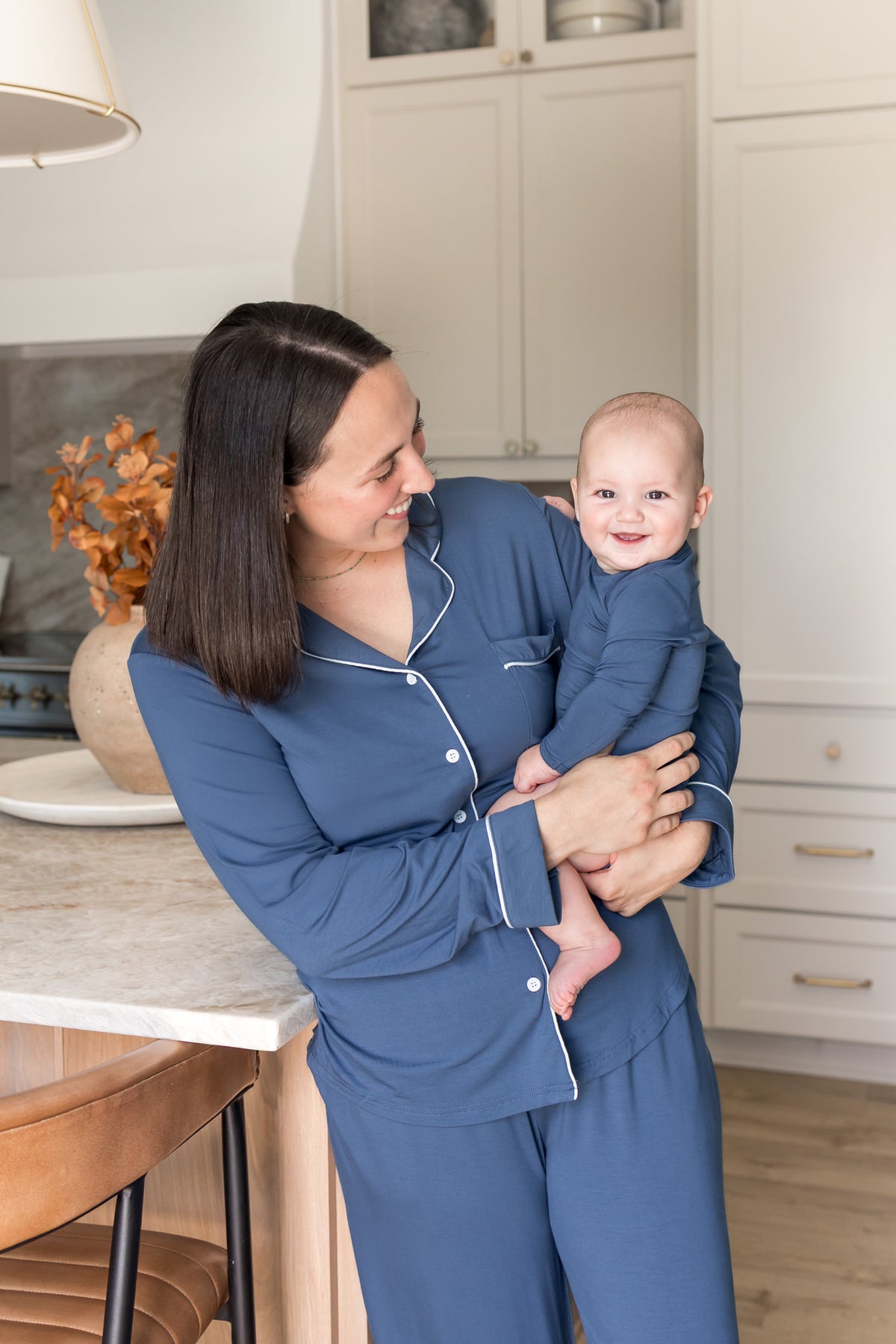 A woman in a navy blue pajama set with white piping holds a baby wearing a matching navy blue outfit in a bright kitchen setting.