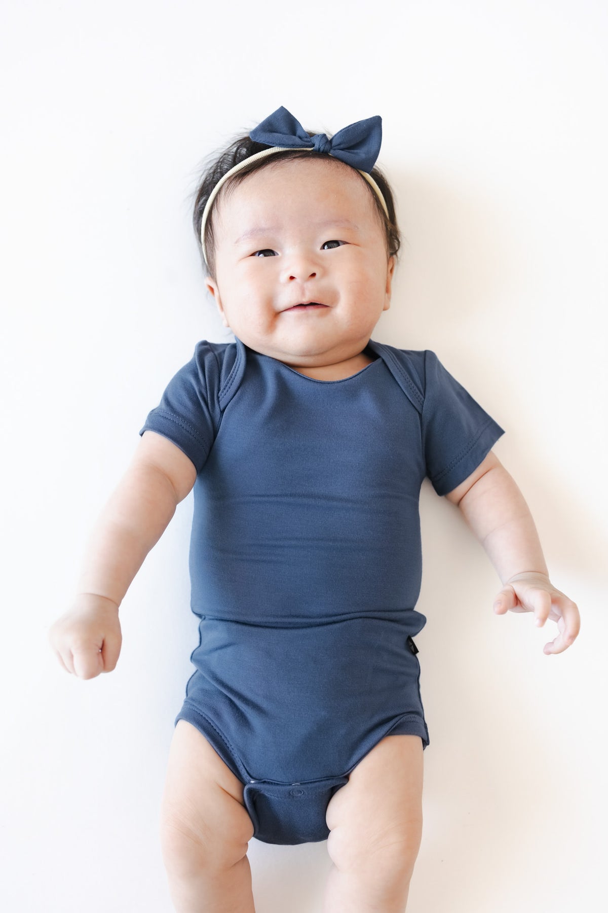 A baby lying on a white background wearing a navy blue short-sleeve bodysuit and Tiny Nylon Bow in Oxford