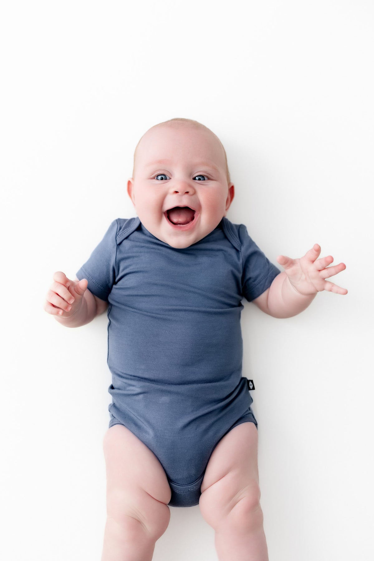 A smiling baby lying on a white background, wearing a short-sleeve blue bodysuit.
