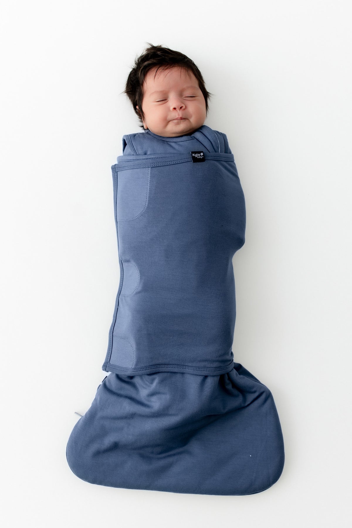 A sleeping newborn baby swaddled in a dusty blue wrap swaddler, lying flat on a clean white background. 