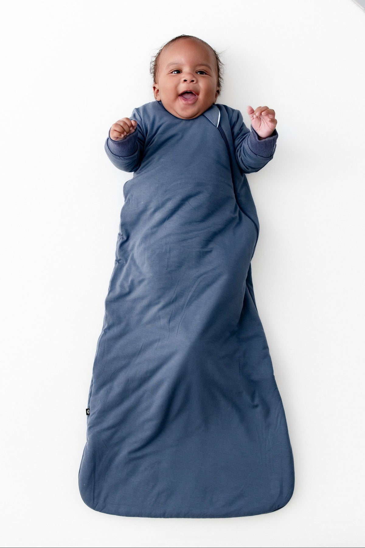 A baby lying on their back, wrapped in a slate blue long sleeve wearable blanket. The baby is smiling and has their hands raised.