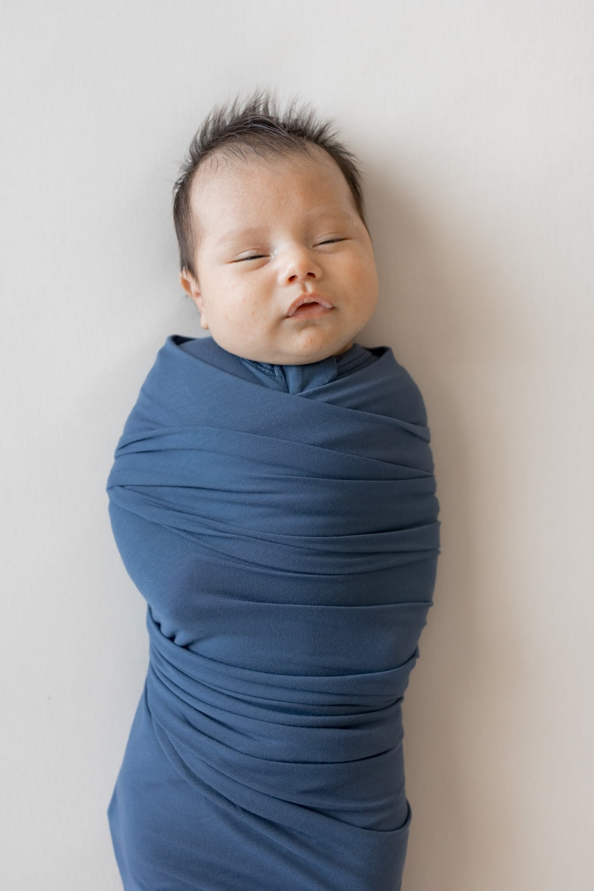 A sleeping newborn baby swaddled in a dusty blue fabric wrap, lying on their side against a plain off-white wall.