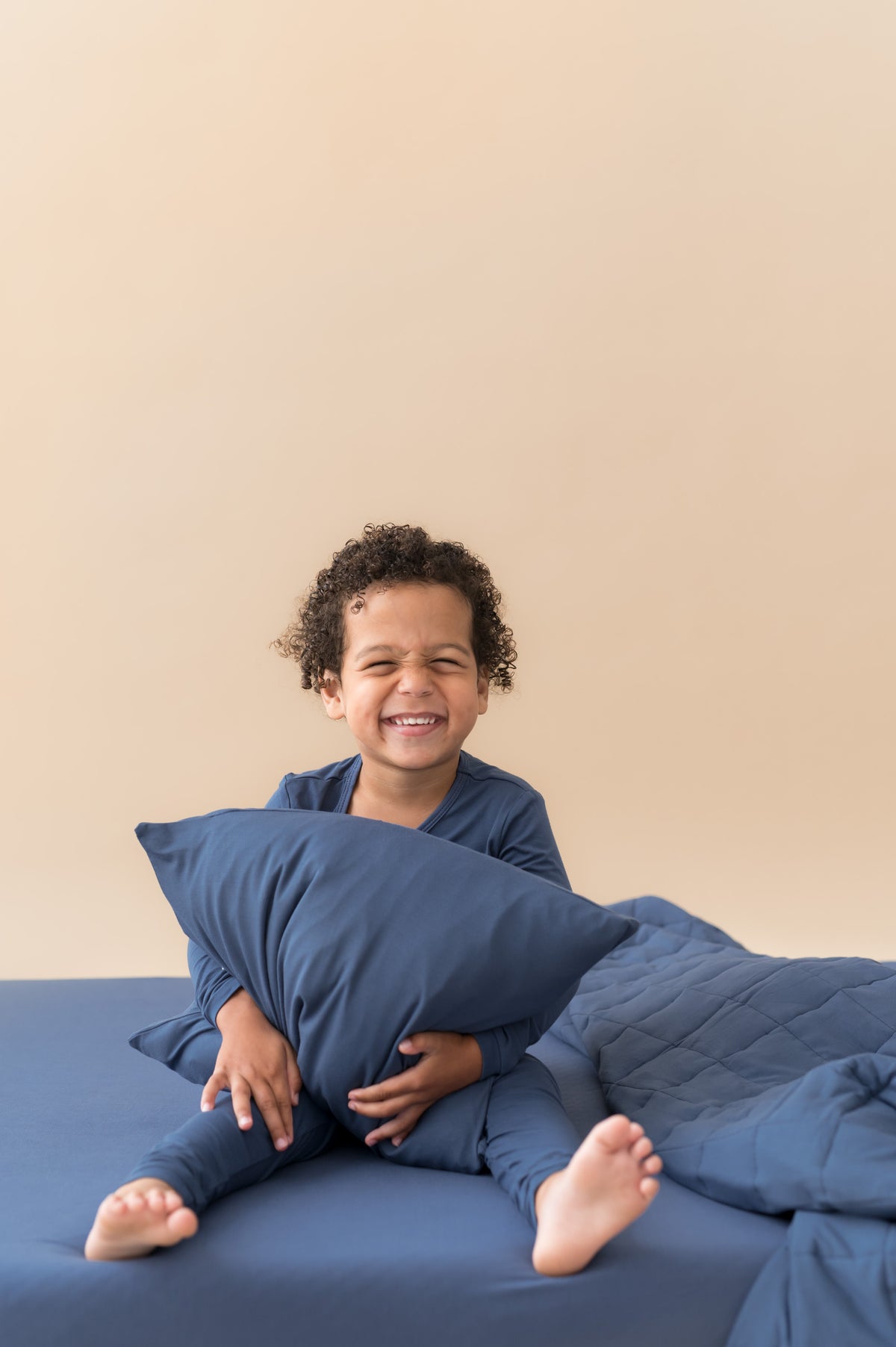 Boy holding Toddler Pillowcase in Oxford sitting with similar color blanket and sheet wearing Oxford long sleeve pajama set