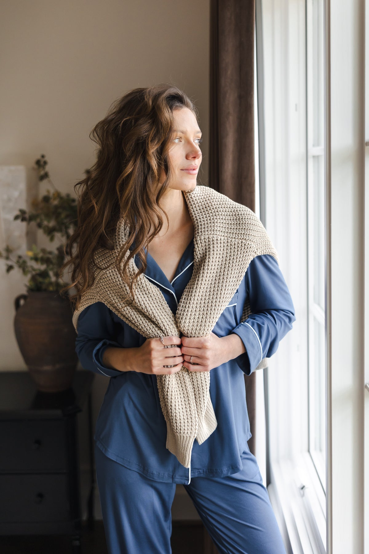 Modal smiling in Long-Sleeved Women's Pajama Set in Oxford with beige sweater over her shoulders looking outside a window