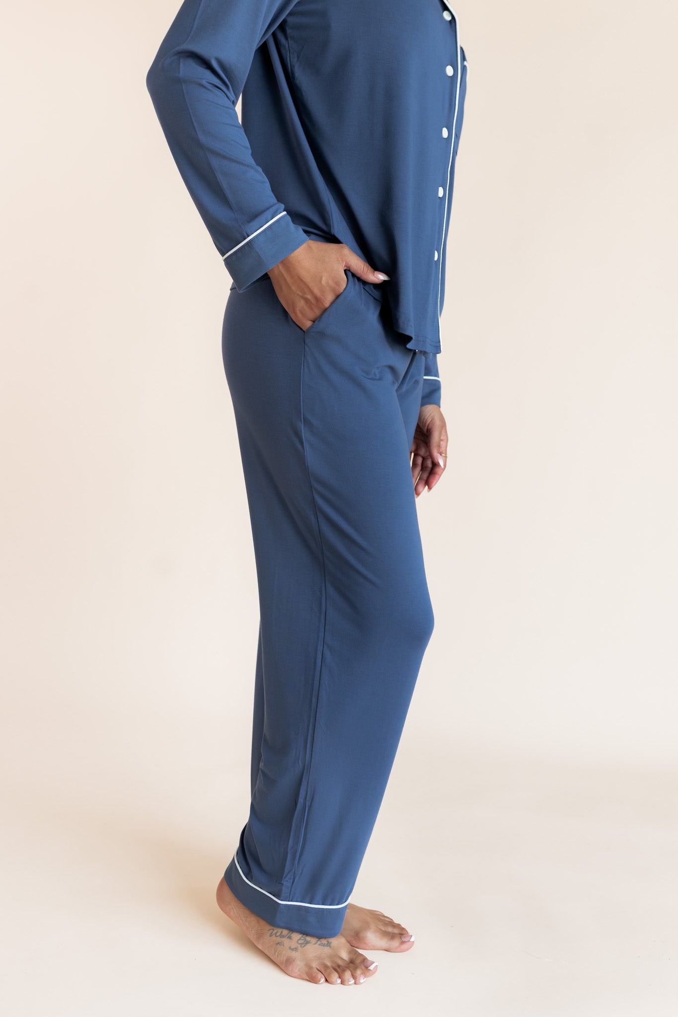 A person wearing a matching two-piece pajama set in slate blue, featuring a long-sleeve button-front top with white piping and matching blue pants with white piping at the cuffs, shown against a plain beige studio background.