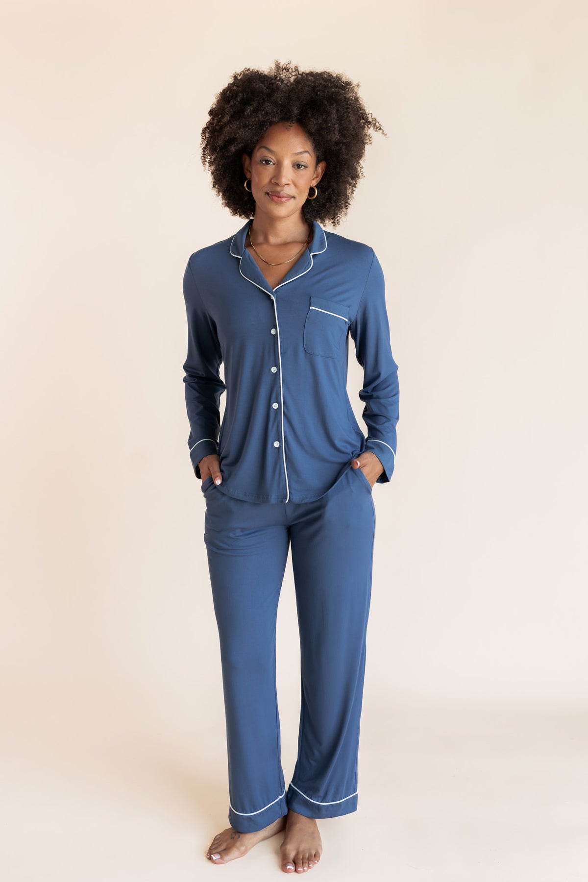 A woman modeling a two-piece blue pajama set with white piping details, consisting of a long-sleeve button-up shirt and matching pants, against a solid light beige background.