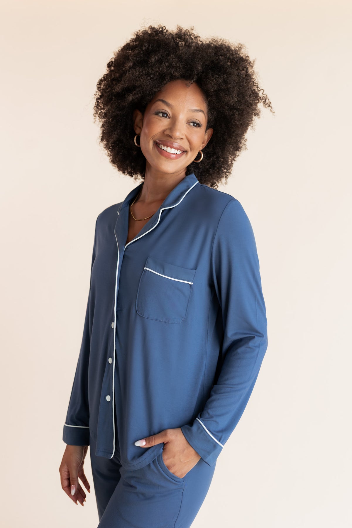 A smiling woman with curly dark hair models a matching blue pajama set with white piping details against a solid light beige background.