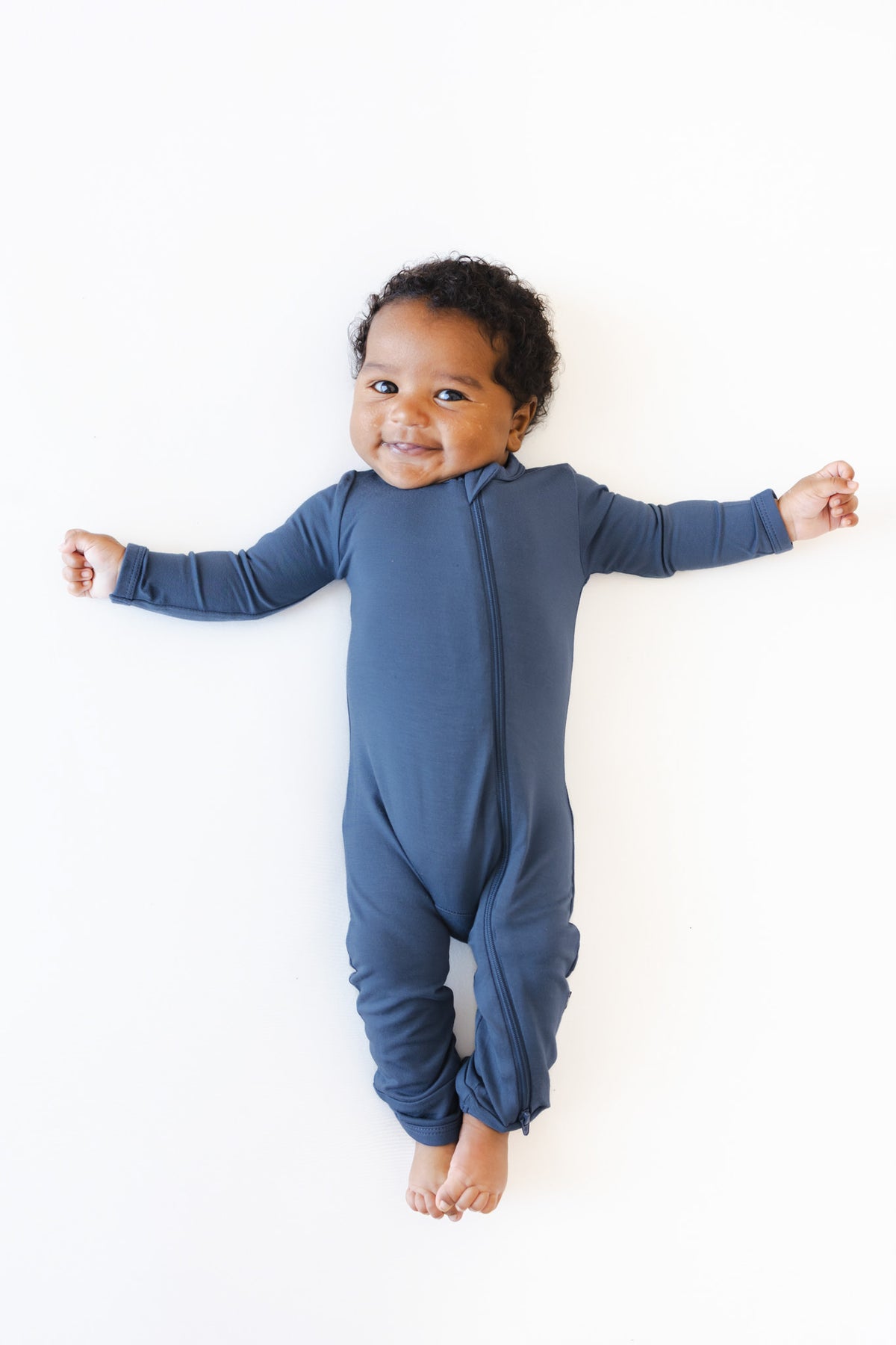 A baby wearing a navy blue long-sleeve footless one-piece romper, lying flat on a white background with arms outstretched.