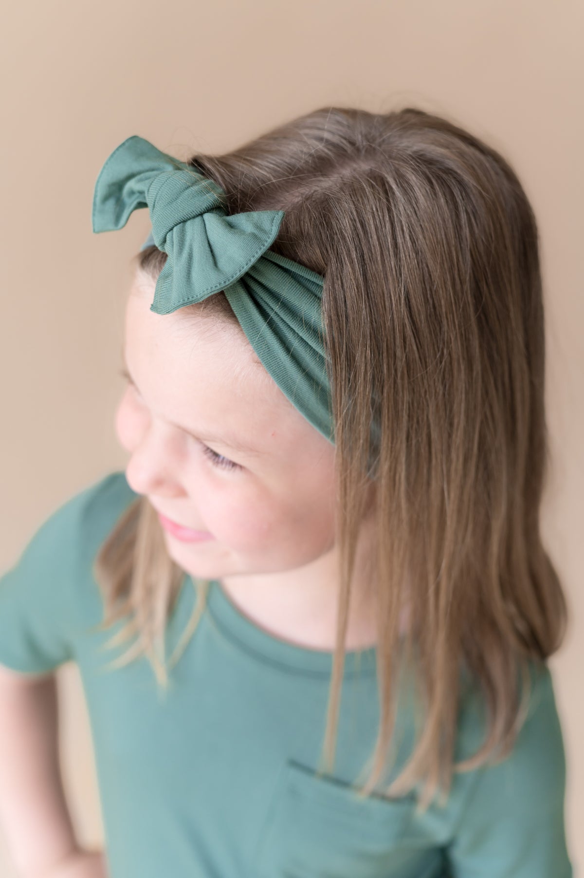 Girl wearing Knotted Bow Headband in Pine over similar color shirt on a beige background
