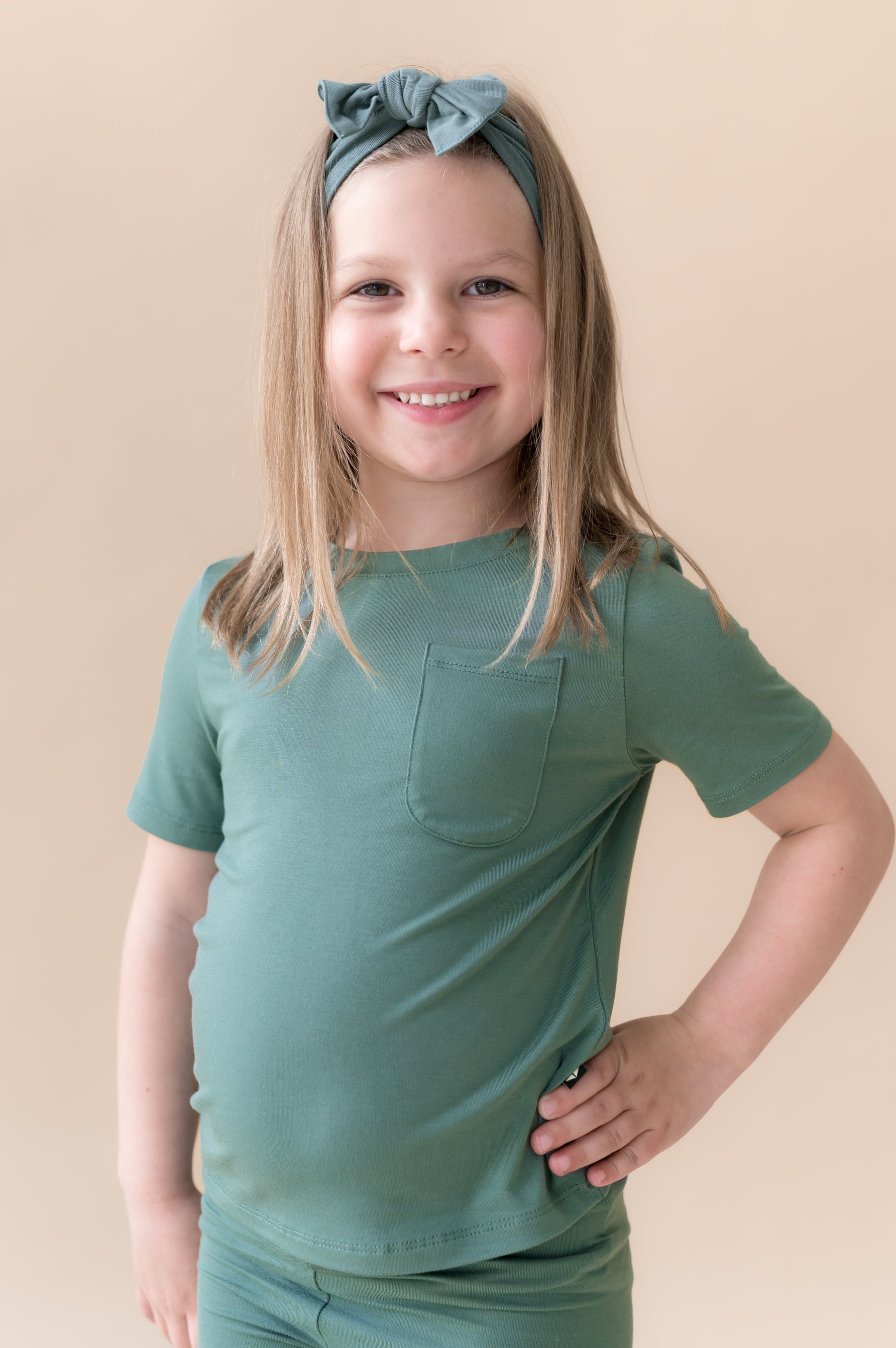 Girl wearing Knotted Bow Headband in Pine over similar color shirt on a beige background