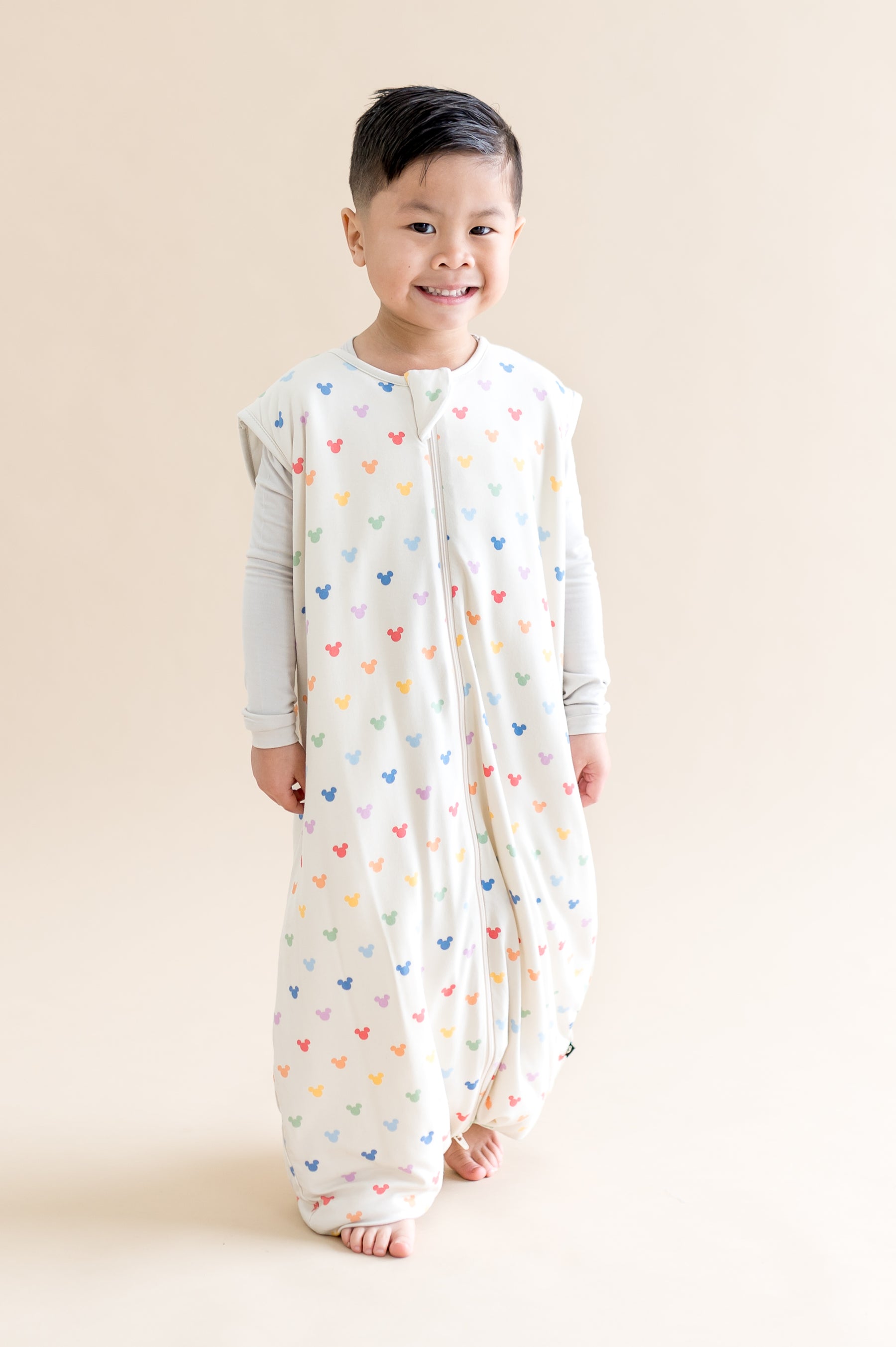 Child wearing a cream sleeveless sleep bag walker with colorful mickey patterns on a beige background