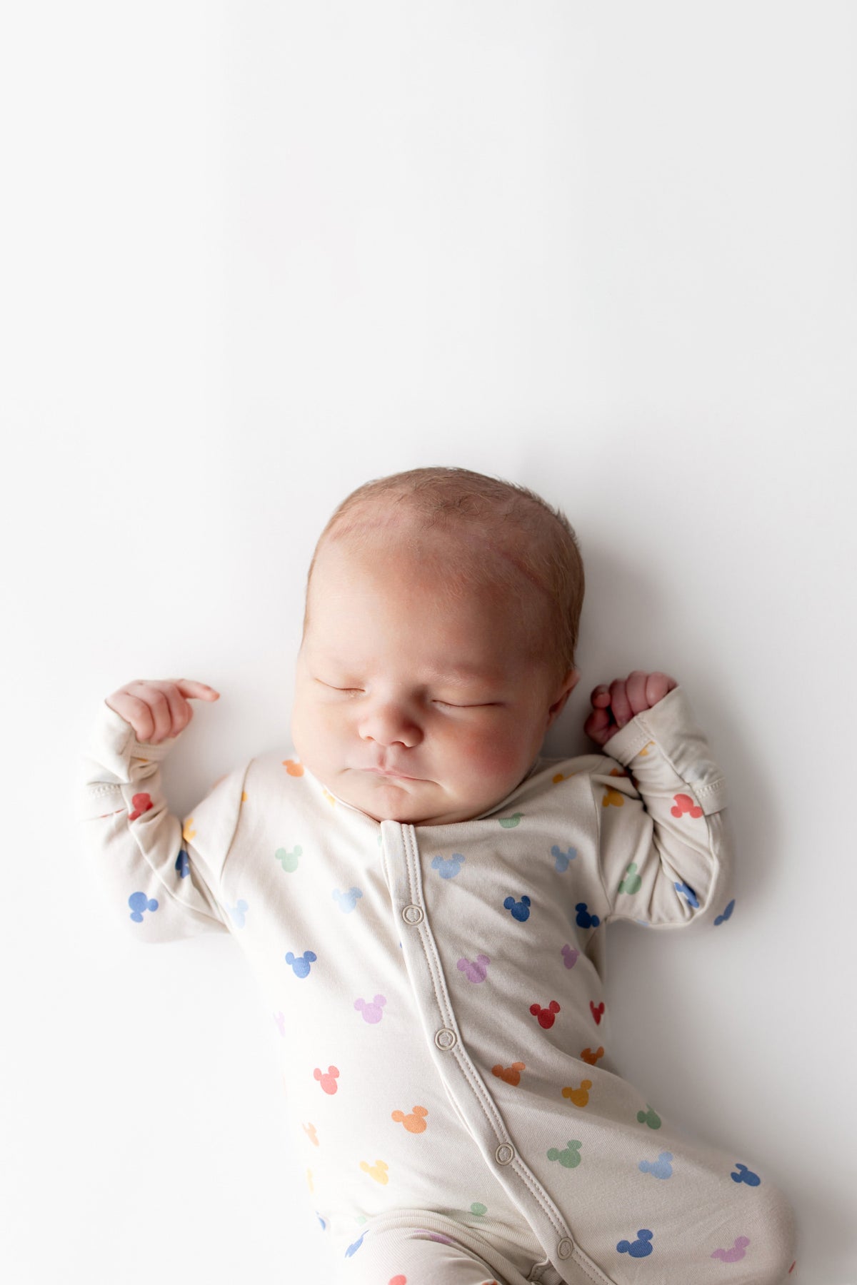 Newborn baby sleeping in a white onesie with colorful mickey patterns on a white background