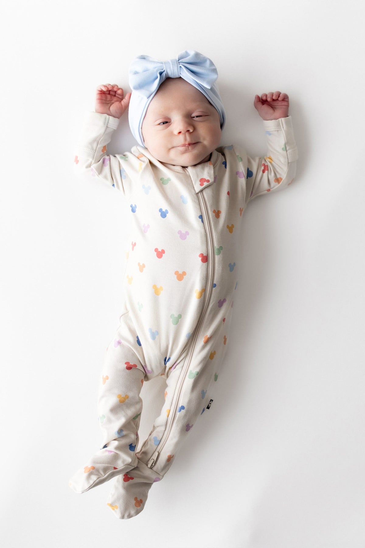 Newborn baby in a colorful mickey footed onesie and blue headband on a white background