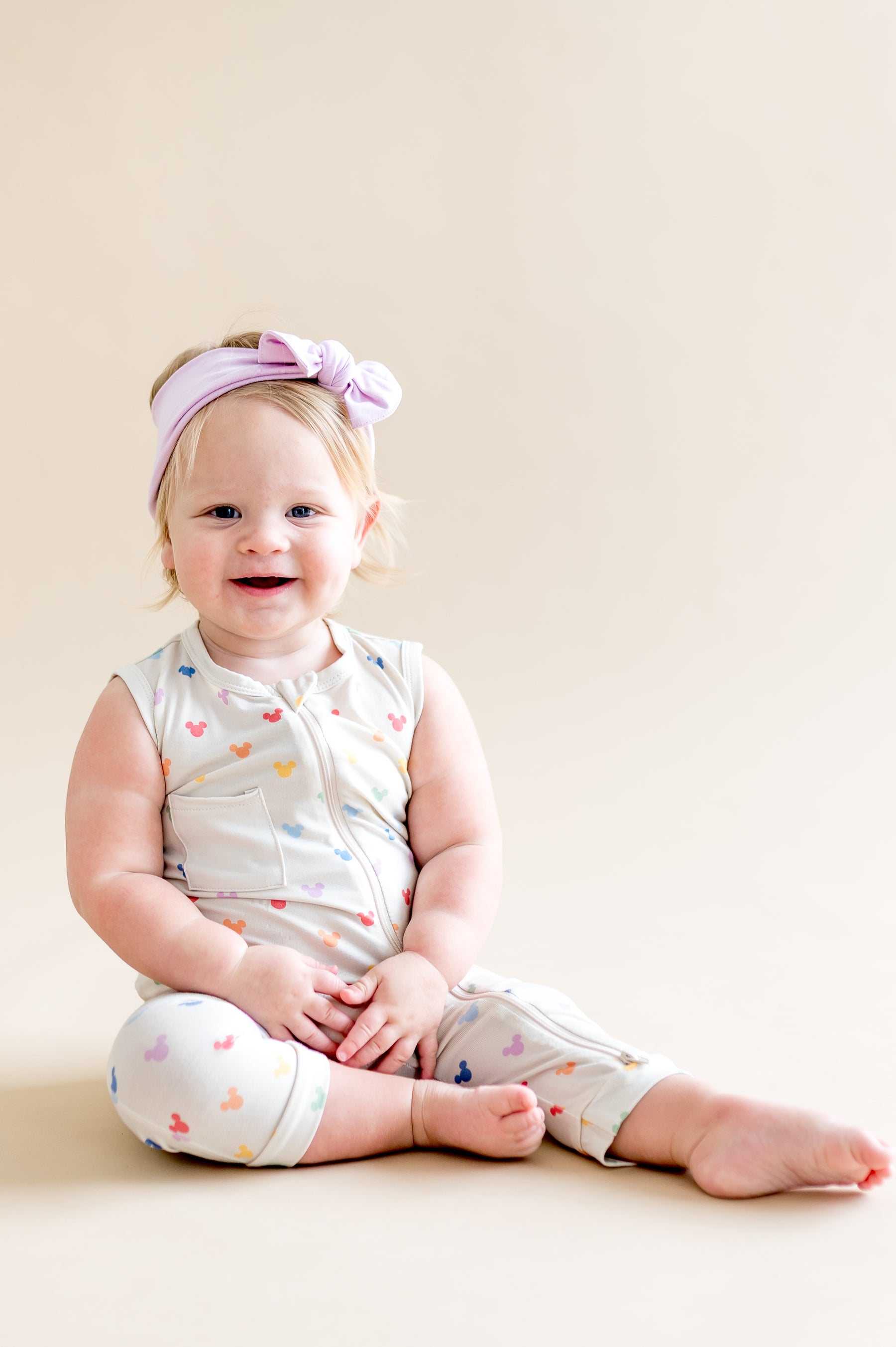 Baby sitting and wearing a zippered sleeveless romper in a colorful mickey head pattern