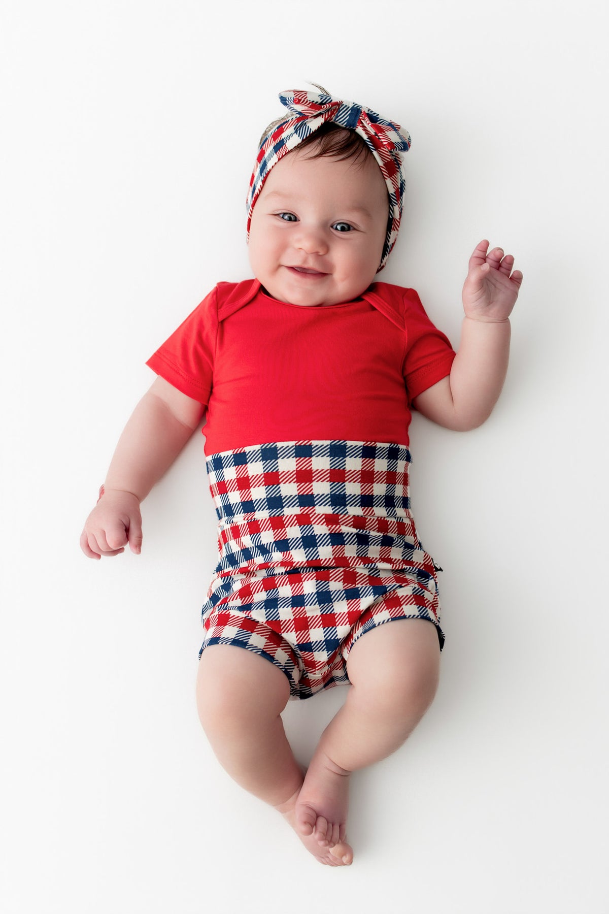 Baby wearing red blue and white Patriotic Gingham check print bummies and similar print bow headband over a cardinal bodysuit on a white background