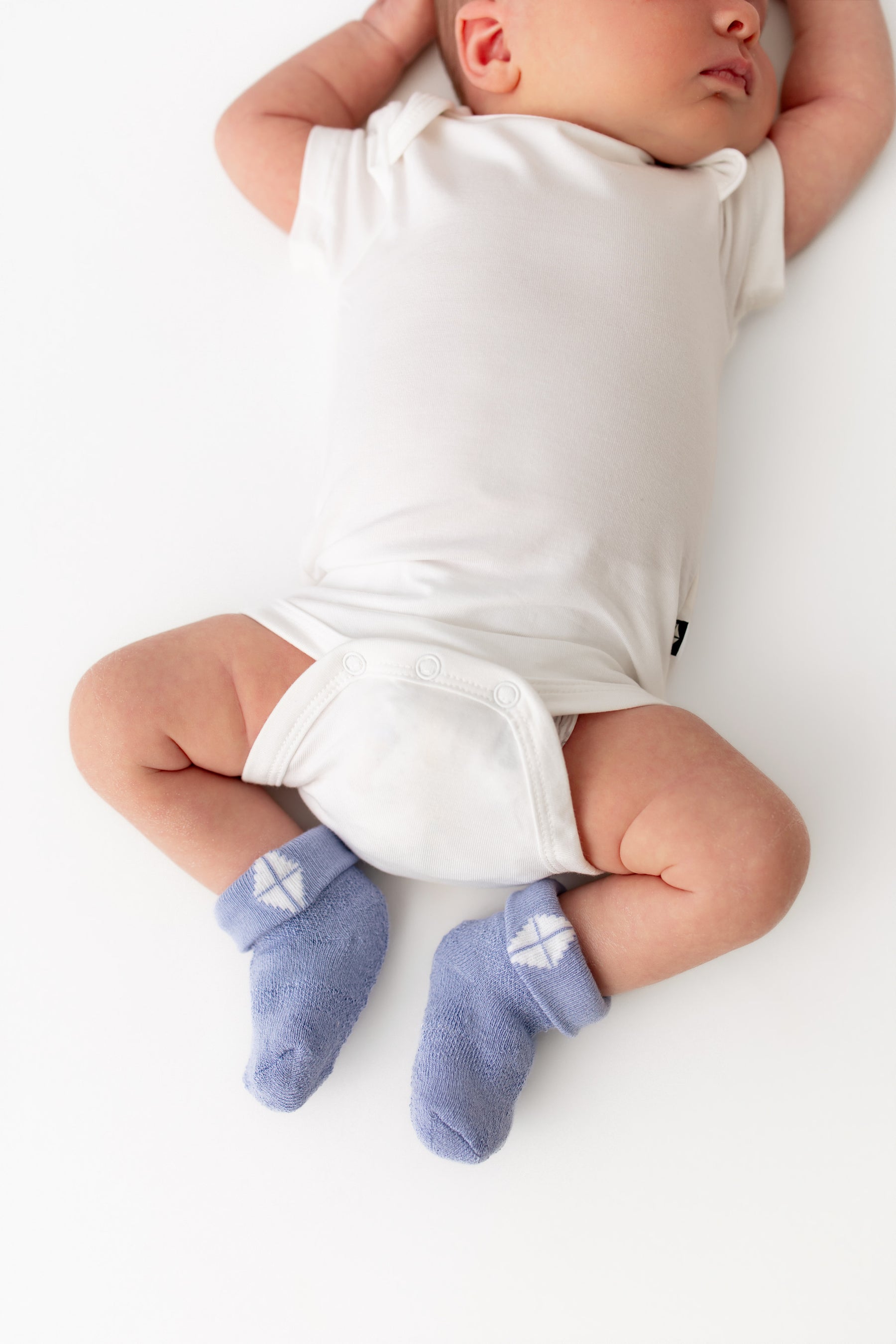 A baby lying on a white surface wearing a white short-sleeve bodysuit and light blue socks with a white geometric pattern.