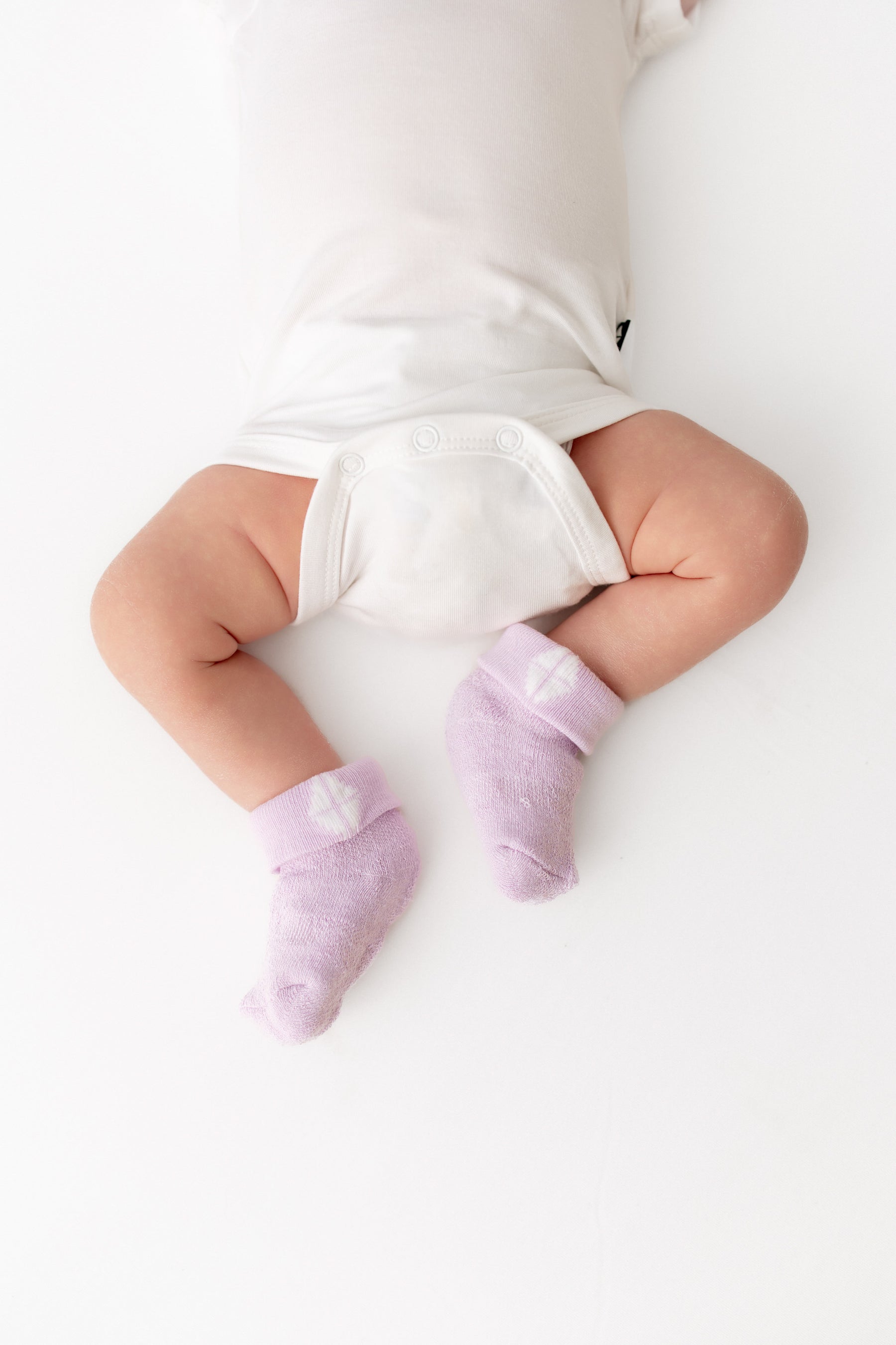 A baby lying on a white surface, wearing light purple knit socks with small white heart details and a white bodysuit.