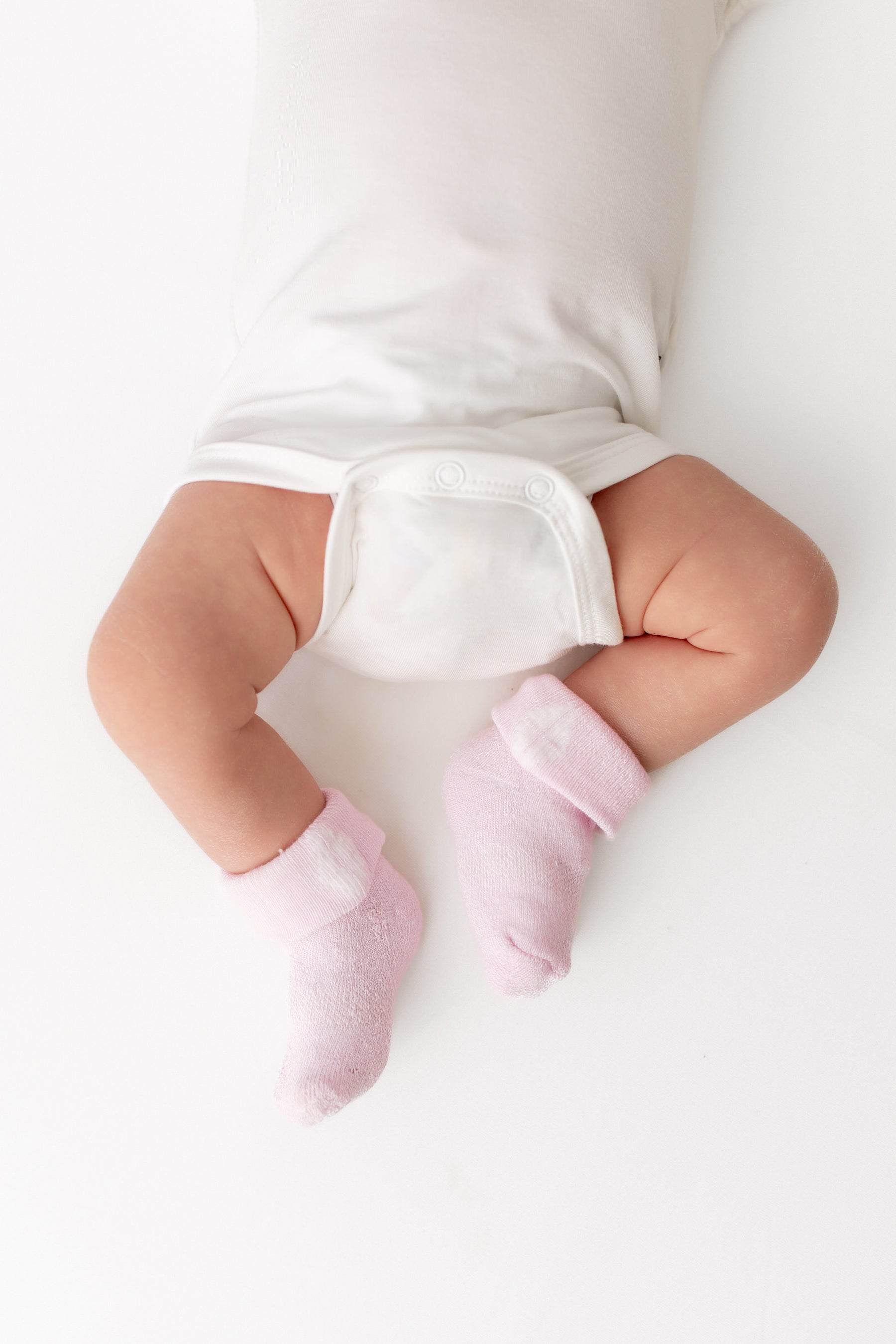 A baby wearing light pink knit booties or socks with a white bodysuit, photographed from above on a clean white background.