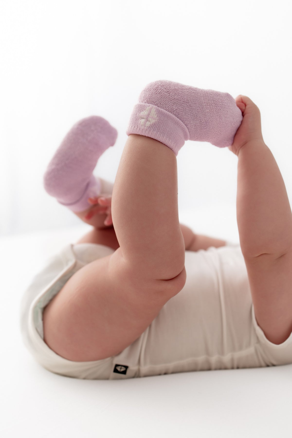 A baby lying on their back wearing light pink knit socks and a cream-colored bodysuit, with their legs raised in the air against a bright white background.