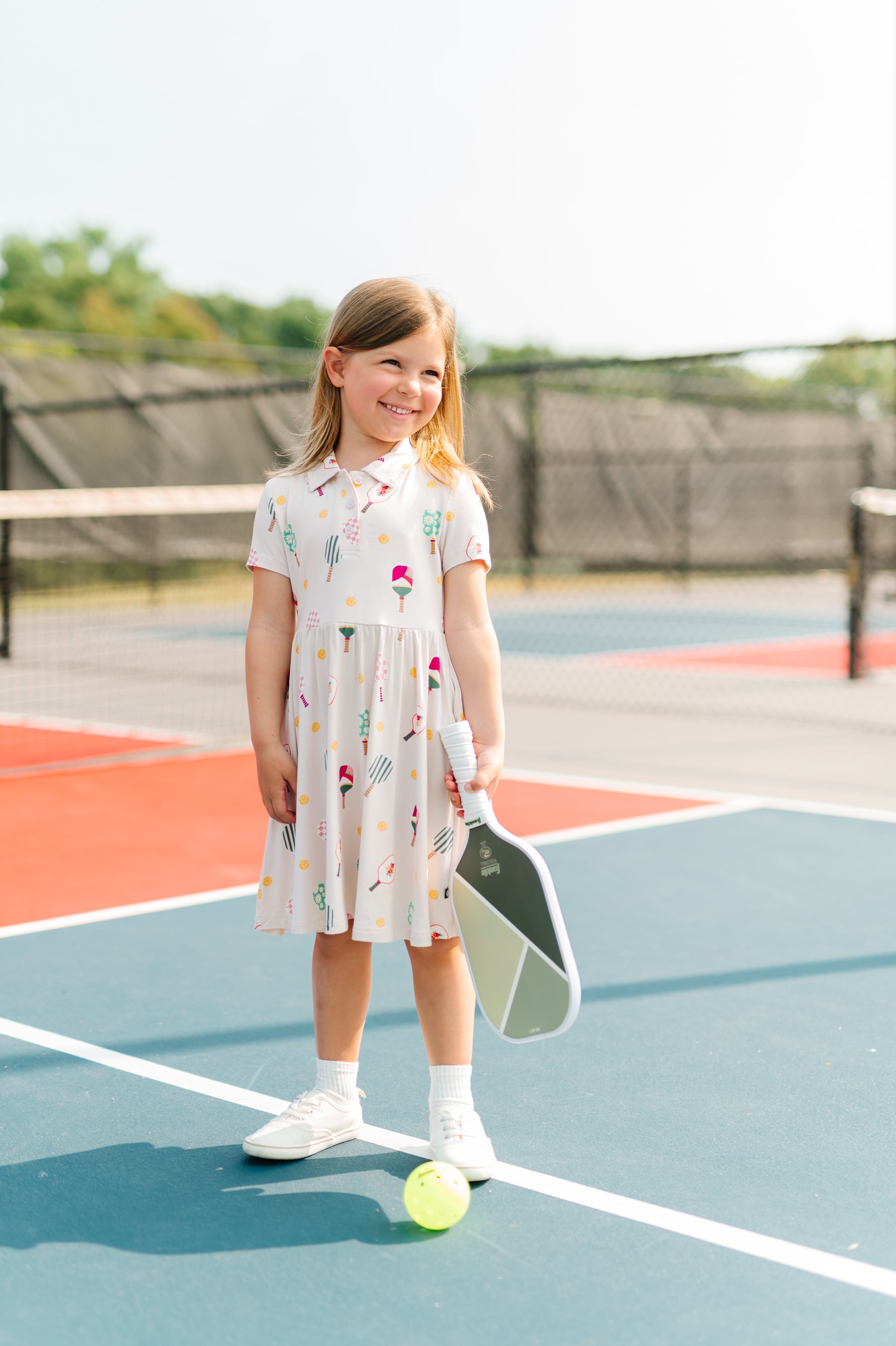 Girl wearing Polo Dress in Pickleball print standing on a pickleball court with bat and ball