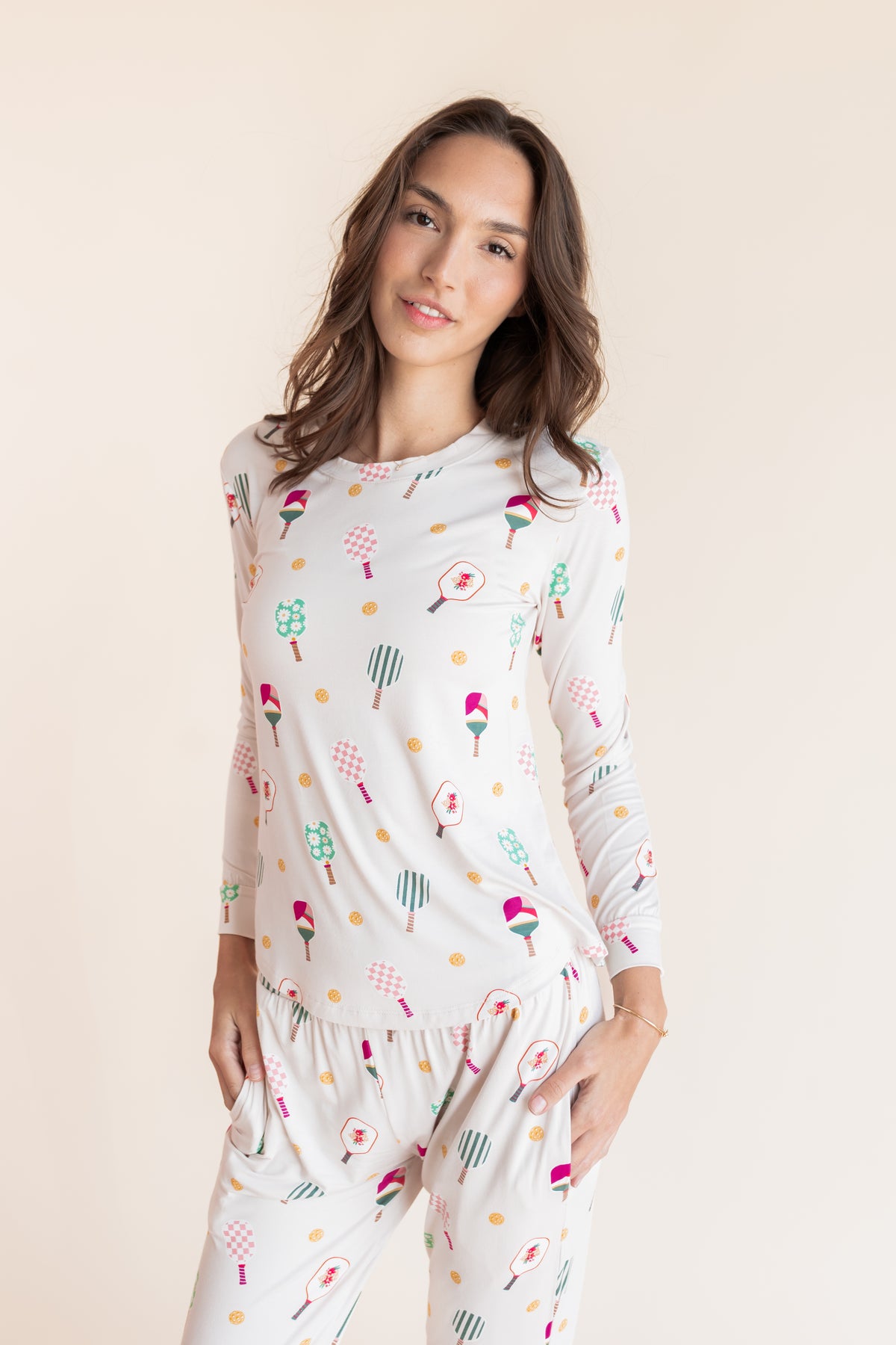 A woman modeling a two-piece pajama set featuring a long-sleeve top and matching pants with a colorful pickleball pattern on a white background, set against a solid light beige backdrop.