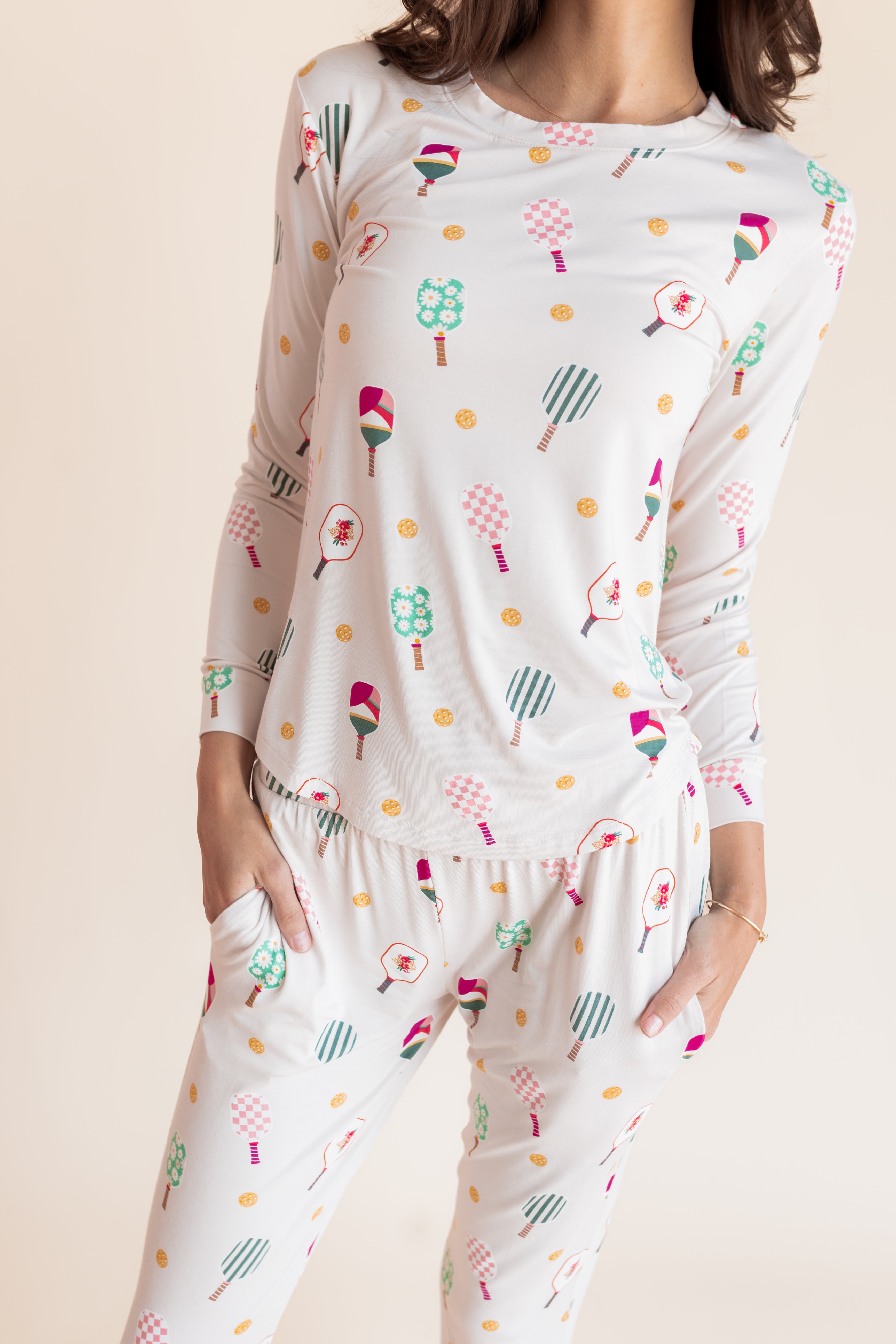 A woman wearing a matching long-sleeve pajama top and pants set with a colorful pickleball print on a white background, photographed against a plain light beige studio backdrop.