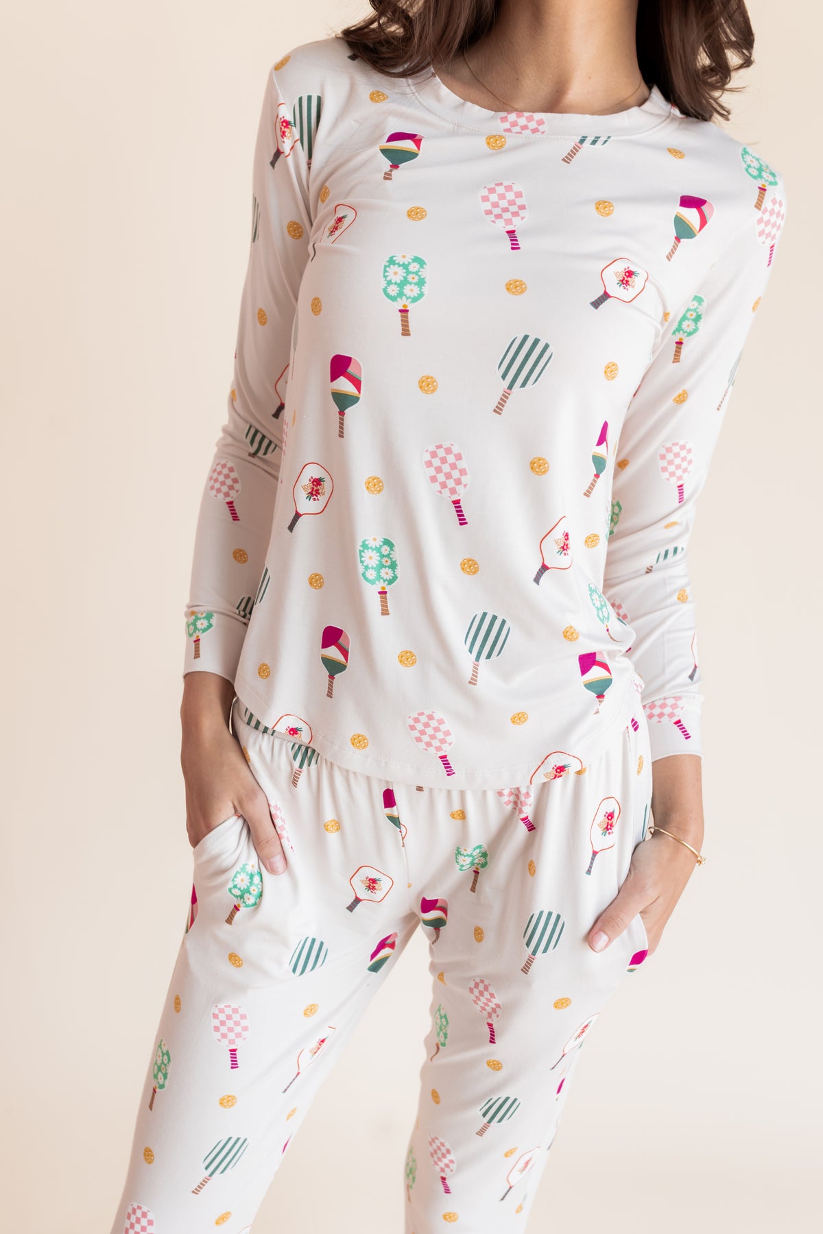 A woman wearing a matching long-sleeve pajama top and pants set with a colorful pickleball print on a white background, photographed against a plain light beige studio backdrop.
